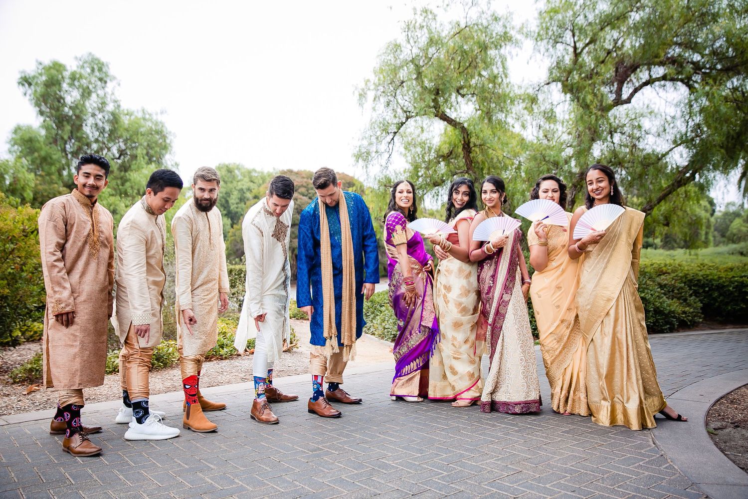 A wedding party in traditional South Asian attire poses together outdoors in a garden setting.