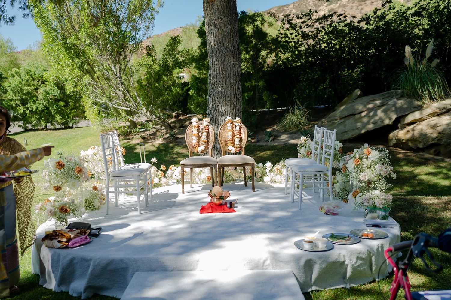 Garden wedding reception area with white chairs and tables arranged under natural tree canopy.