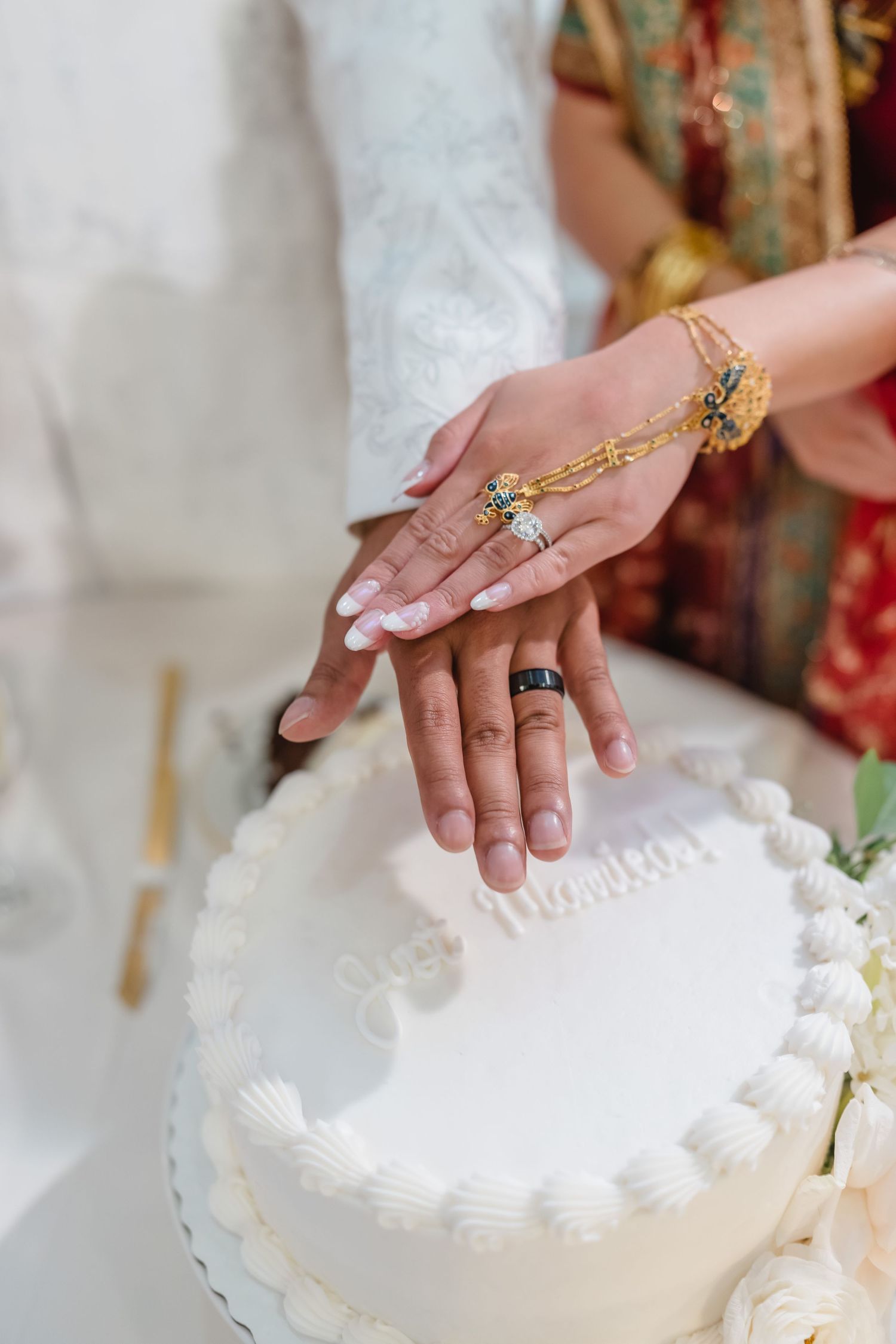 Close-up of hands with ornate jewelry cutting a white wedding cake together.