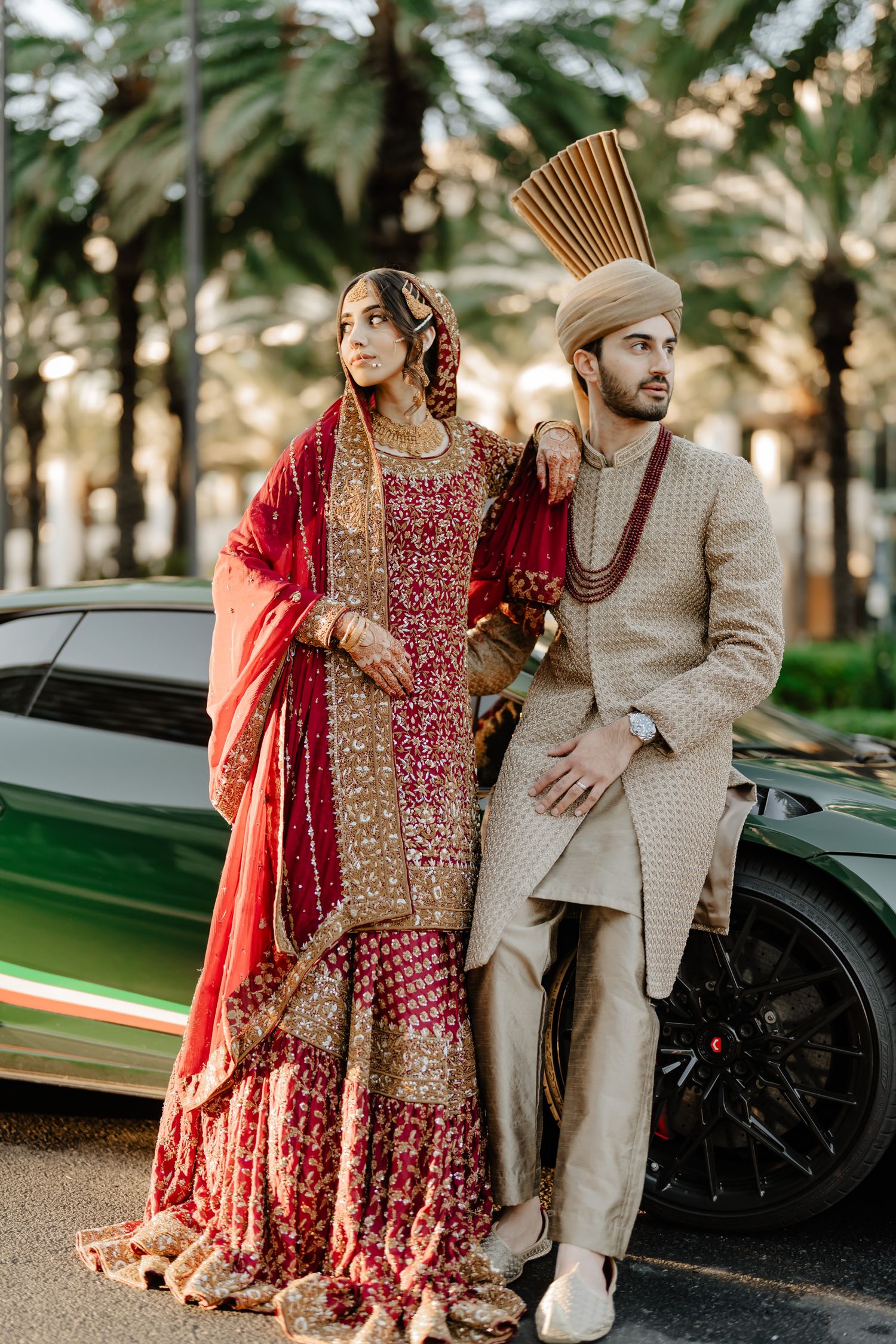 A couple in traditional South Asian wedding attire poses in front of palm trees and a luxury car.
