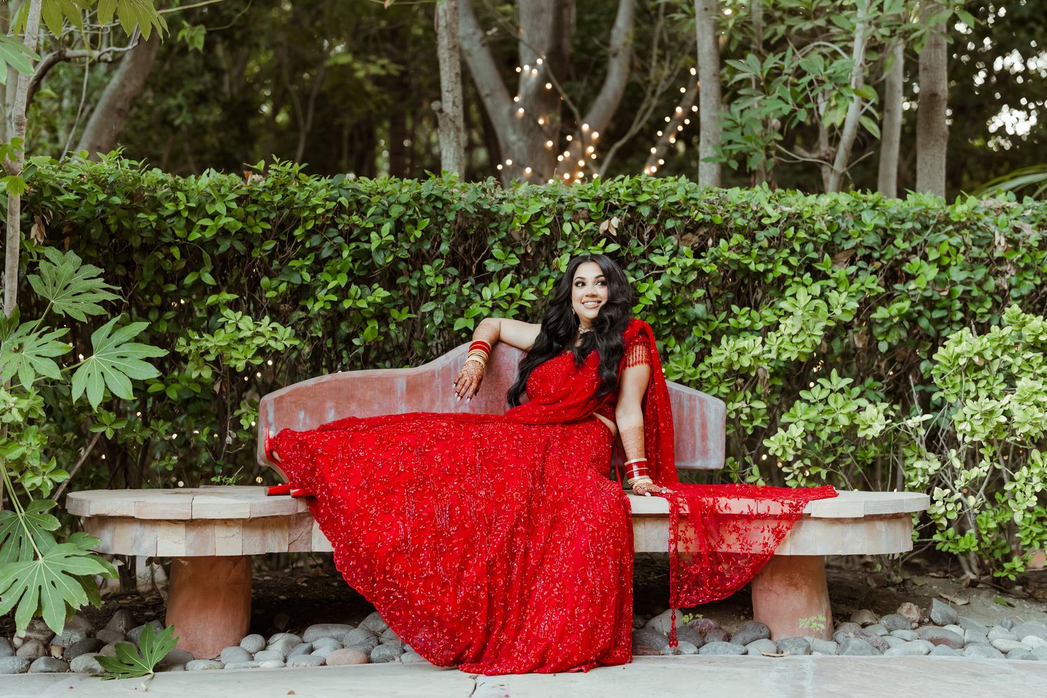 A red sequined dress flows elegantly across a garden bench surrounded by tropical foliage.
