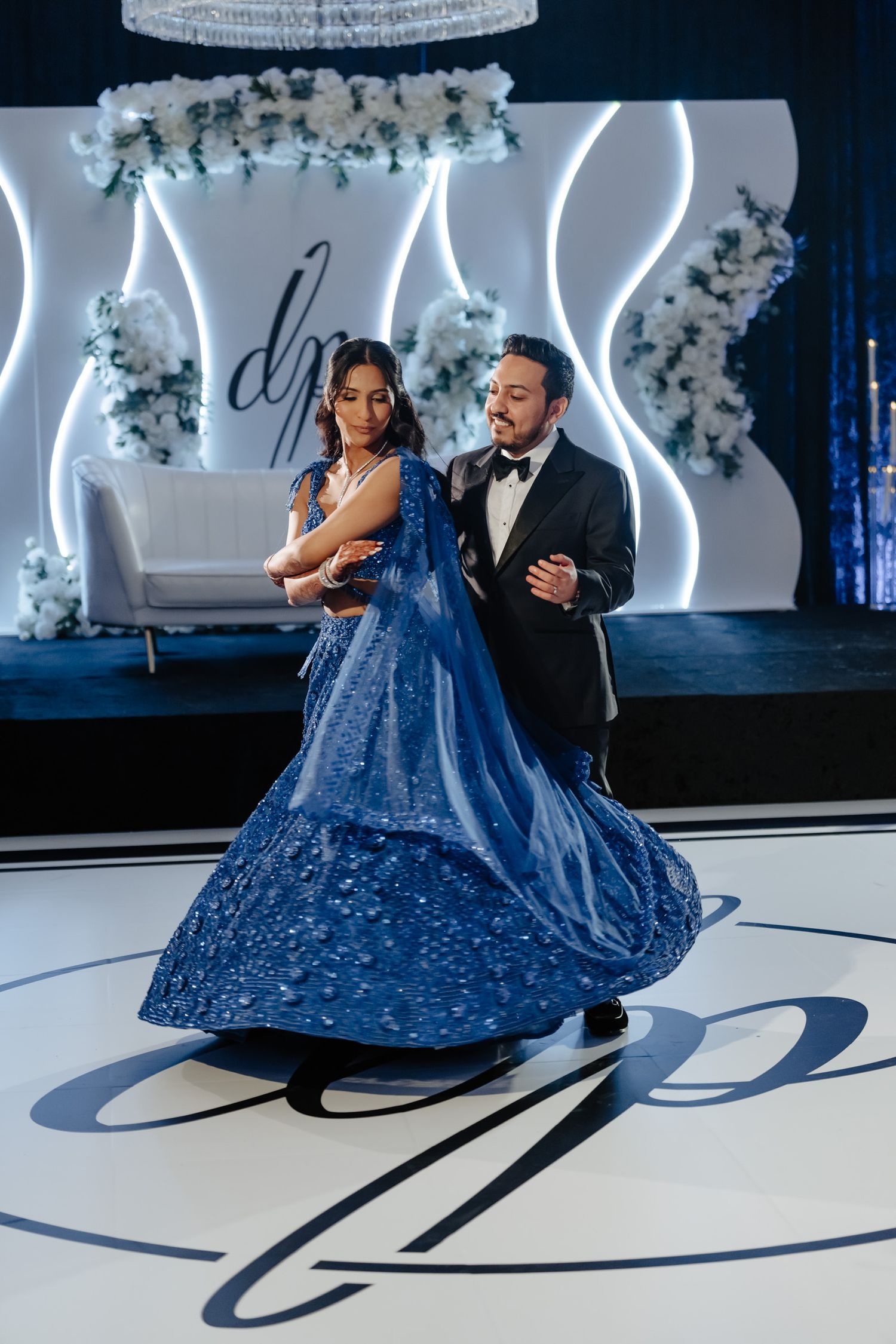A couple dances elegantly on a monogrammed dance floor with blue lighting and floral decorations.
