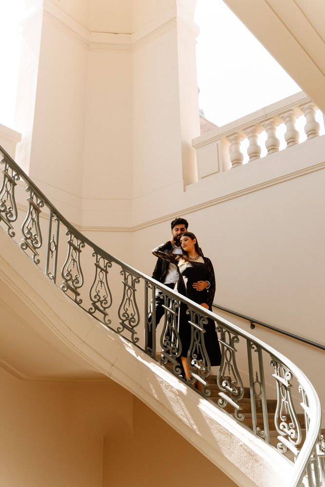 Couple posing romantically on an elegant curved staircase with ornate iron railings in a white building.