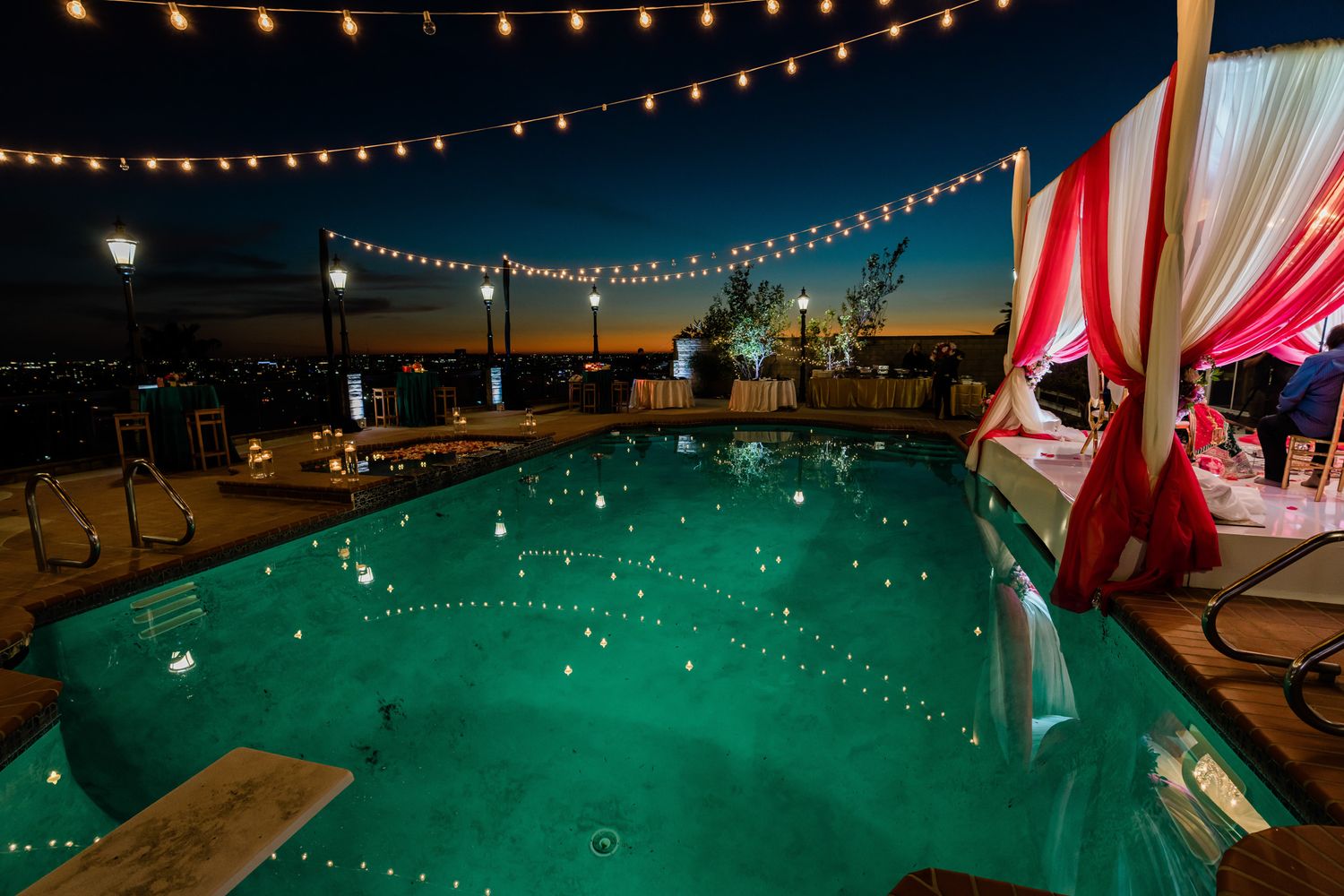 Evening poolside wedding reception setup with twinkling string lights and pink fabric draping.
