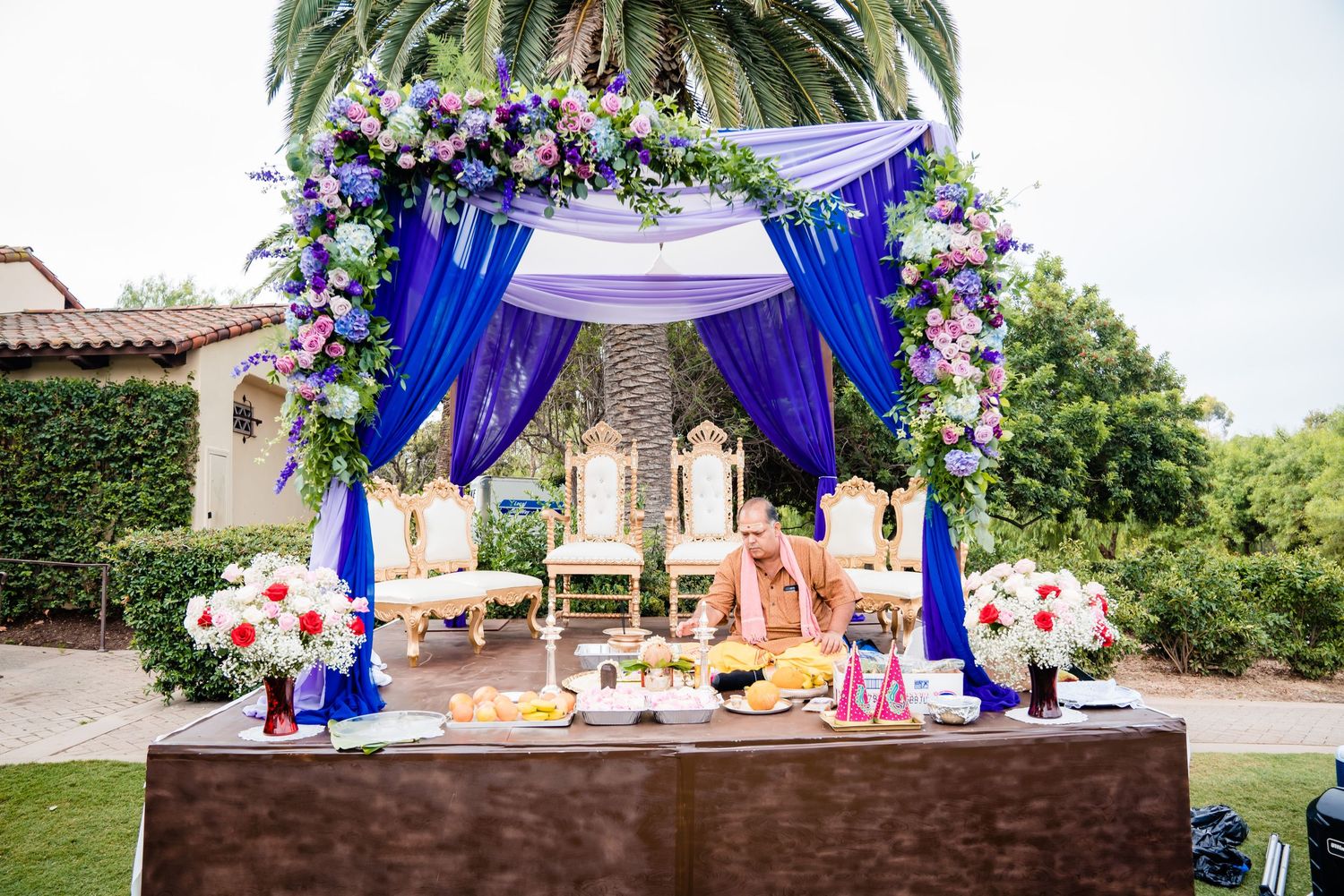 Elegant outdoor wedding ceremony setup with royal blue drapes and white floral arrangements under a palm tree canopy.