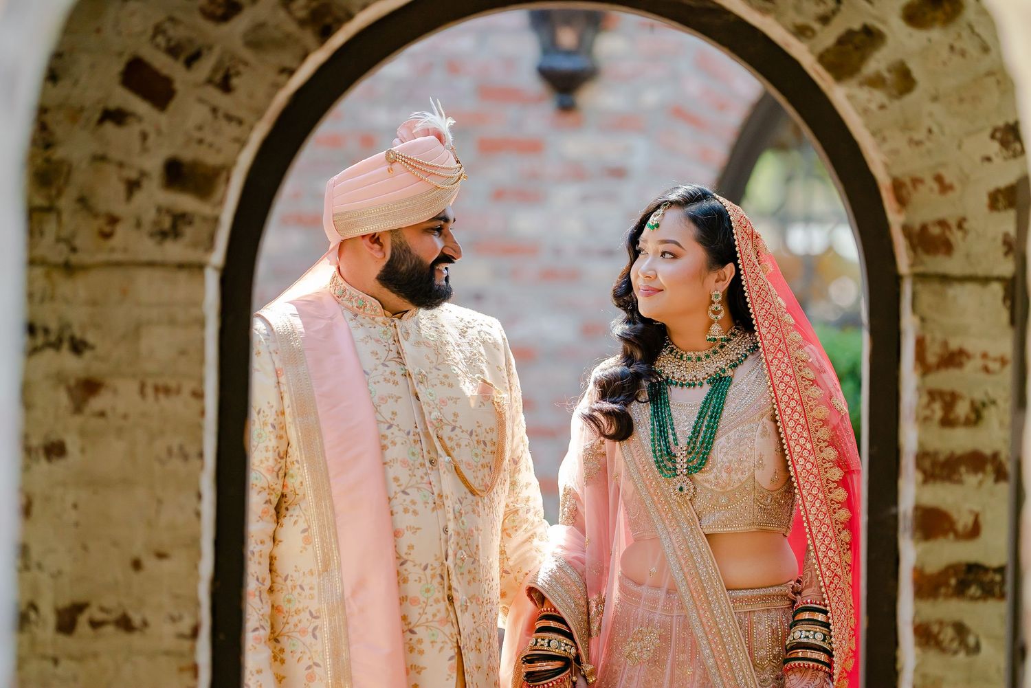 A couple in traditional Indian wedding attire share romantic moments under stone archways during their celebration.
