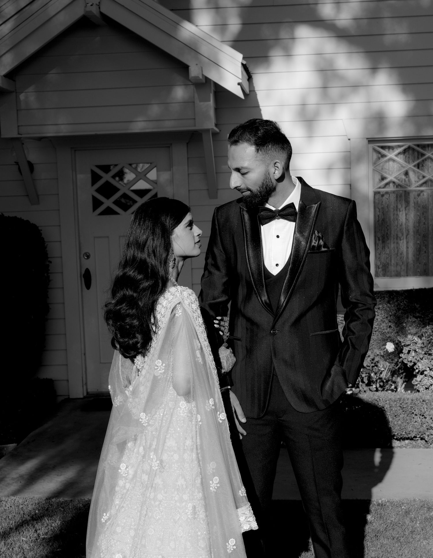 Romantic black and white portrait of a couple in wedding attire standing by a rustic building.