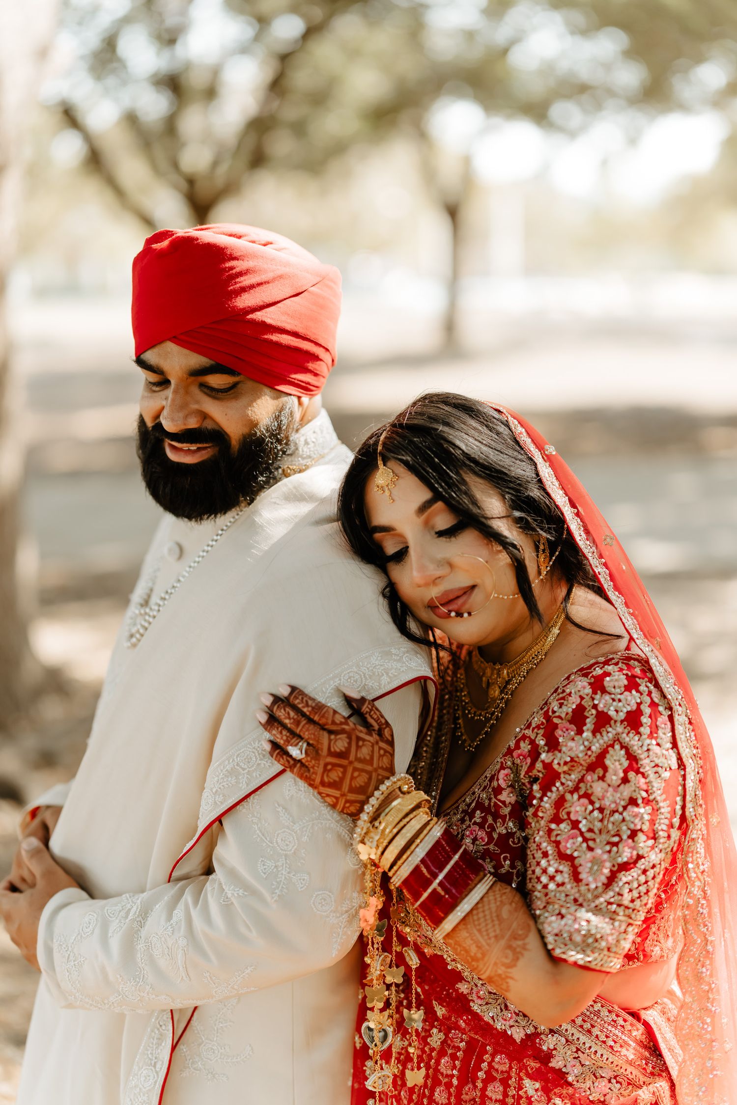 A couple in traditional Sikh wedding attire embrace while the bride admires her mehndi decorations in soft natural lighting.