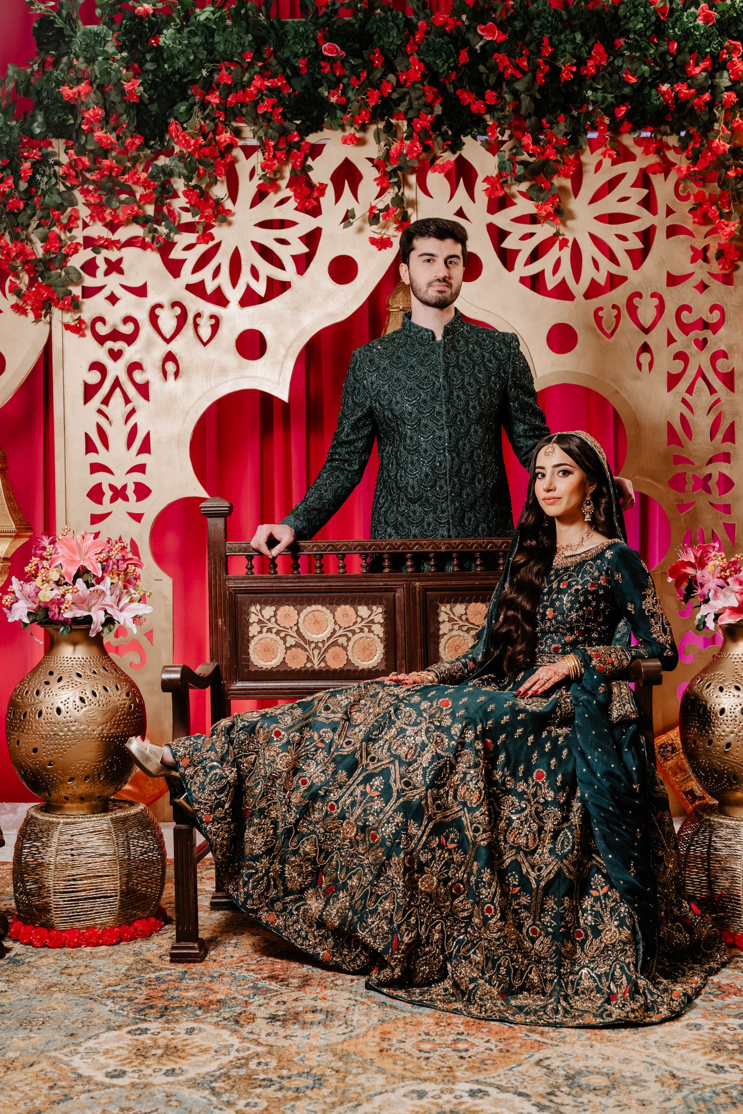 Elegant couple in traditional South Asian attire pose against ornate red and white backdrop with decorative vases.