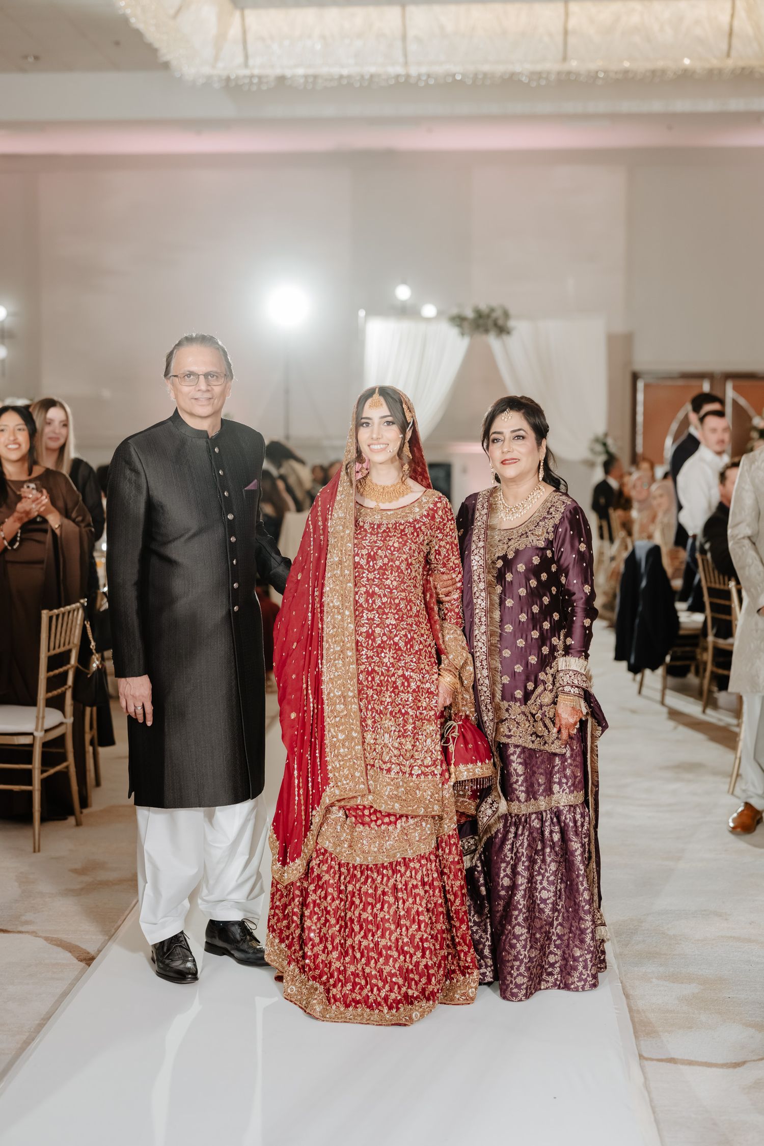 Three people in traditional South Asian wedding attire pose together at an indoor celebration venue.