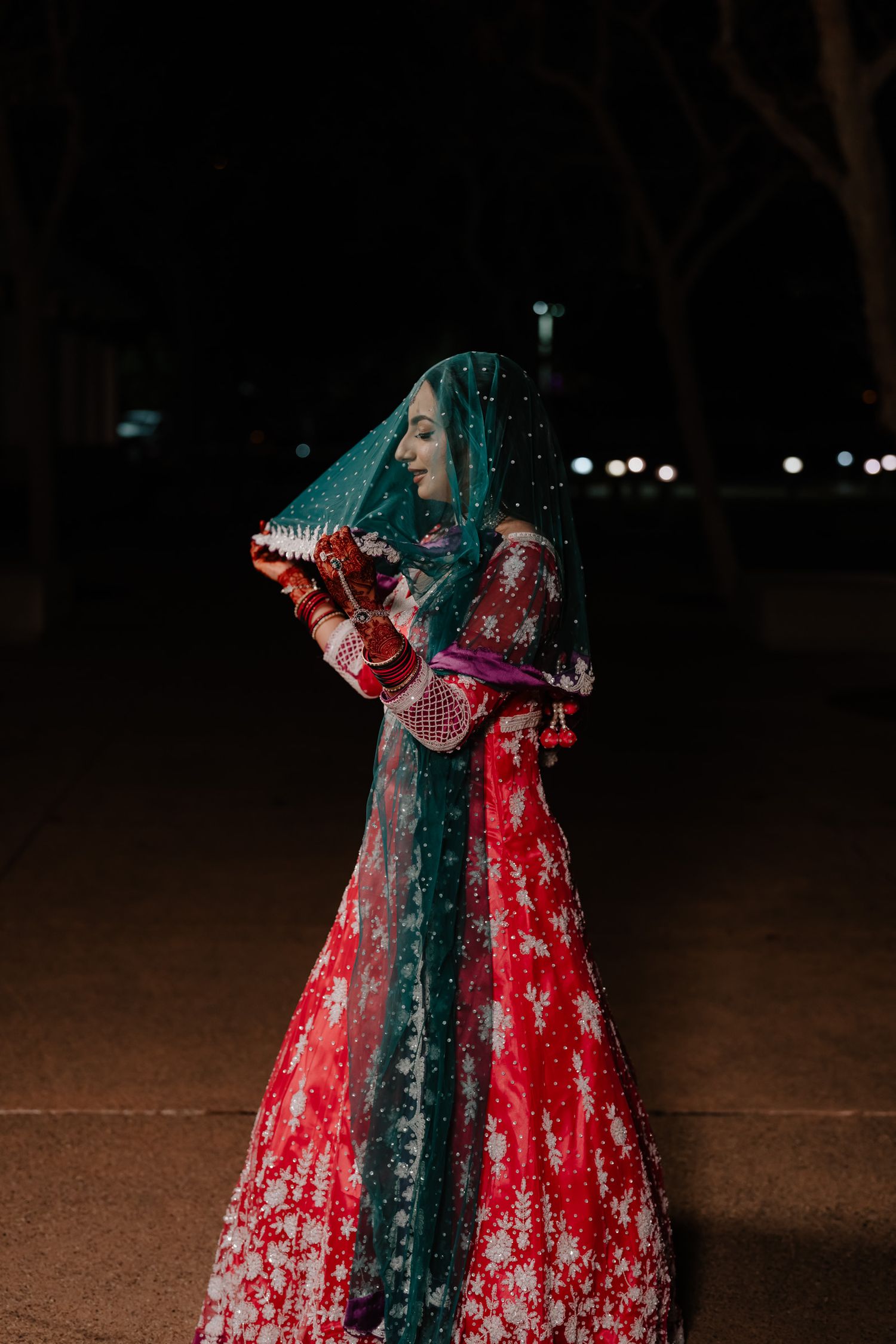 Dramatic nighttime portrait of a bride in a red embroidered lehenga with a teal veil.