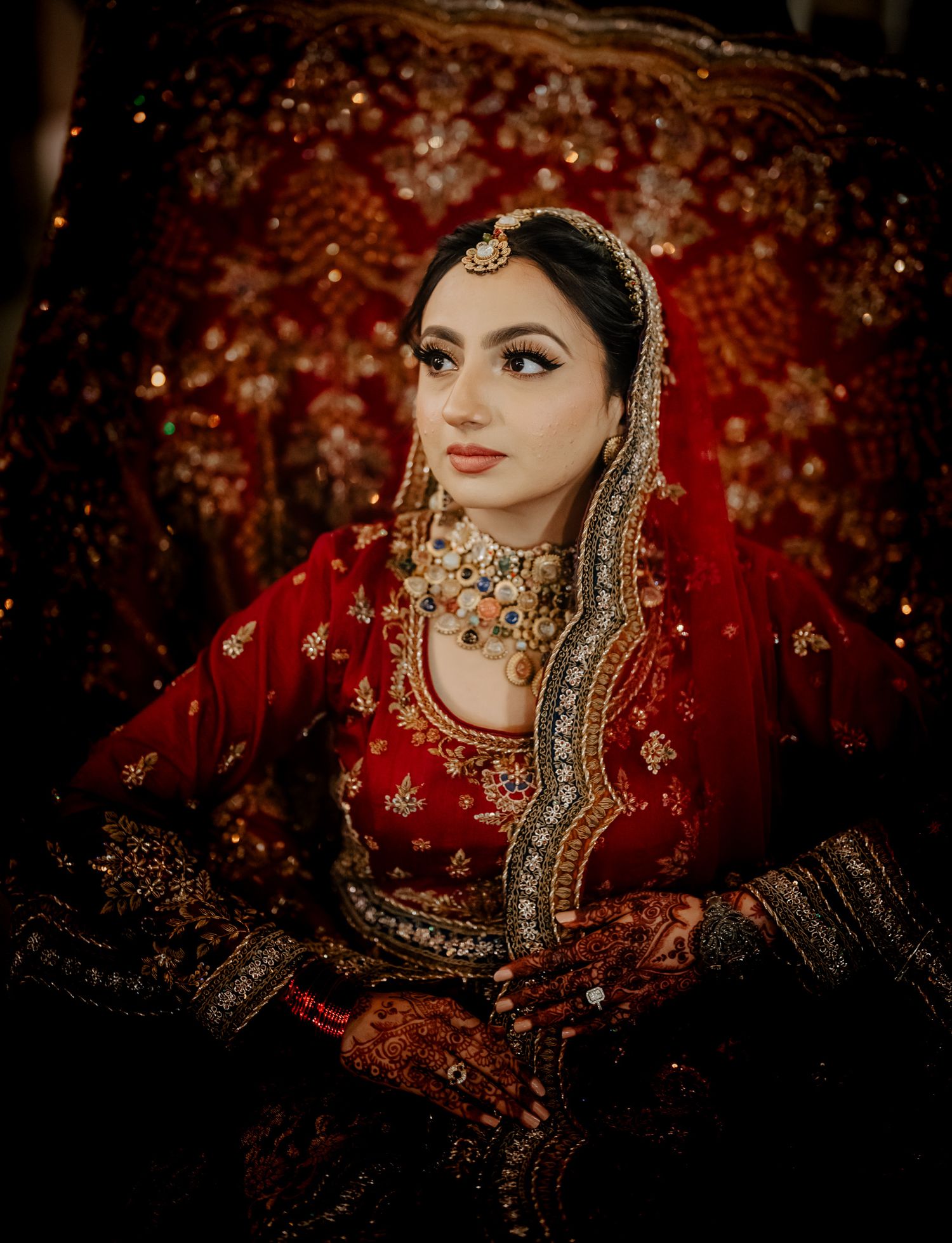 Traditional Indian bride wearing ornate red lehenga and gold jewelry against a bokeh autumn background.