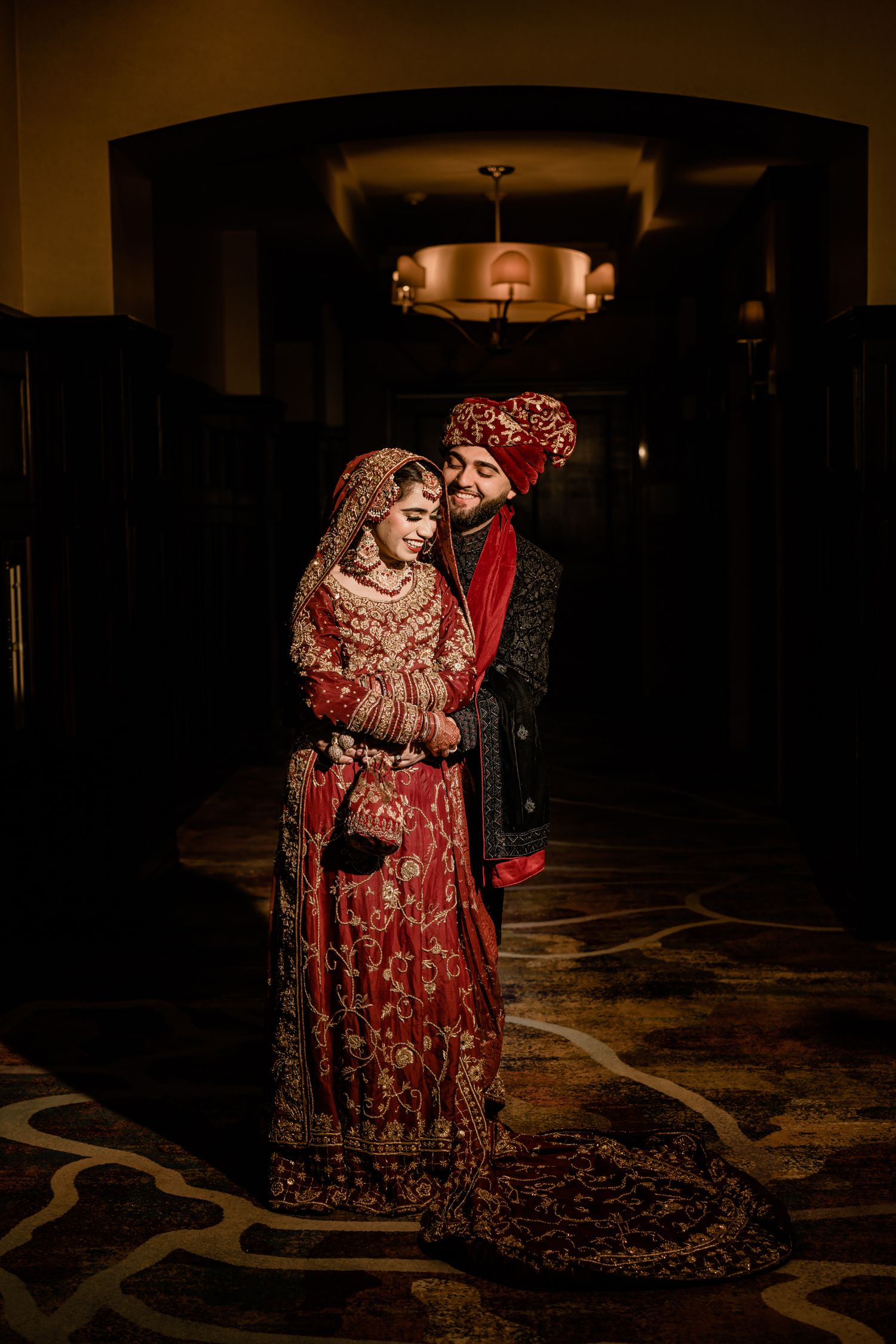 A couple in traditional Indian wedding attire pose together in a dimly lit venue with ornate carpet patterns.