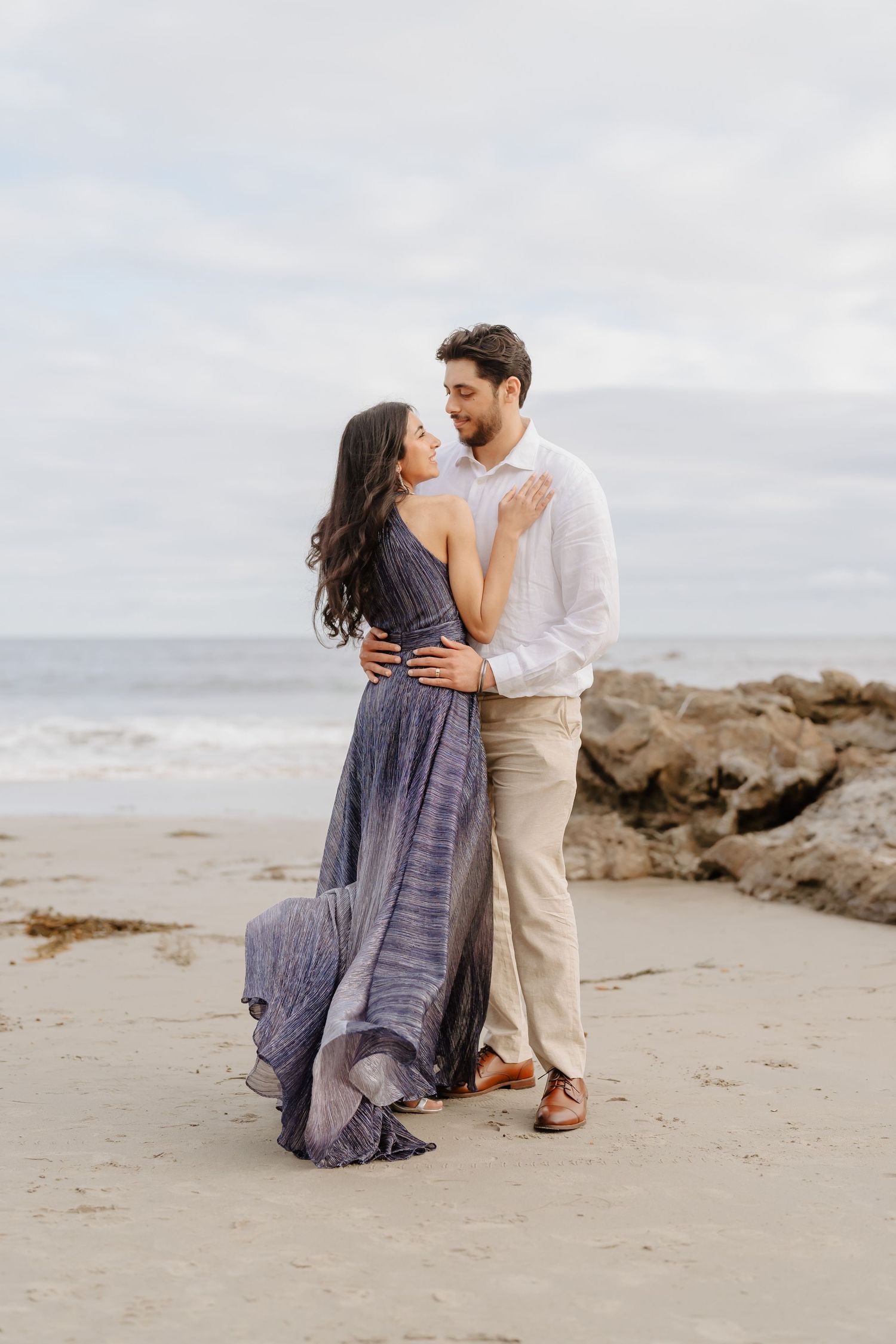 A couple embraces romantically on a sandy beach, woman in flowing blue dress, man in white shirt and khaki pants.