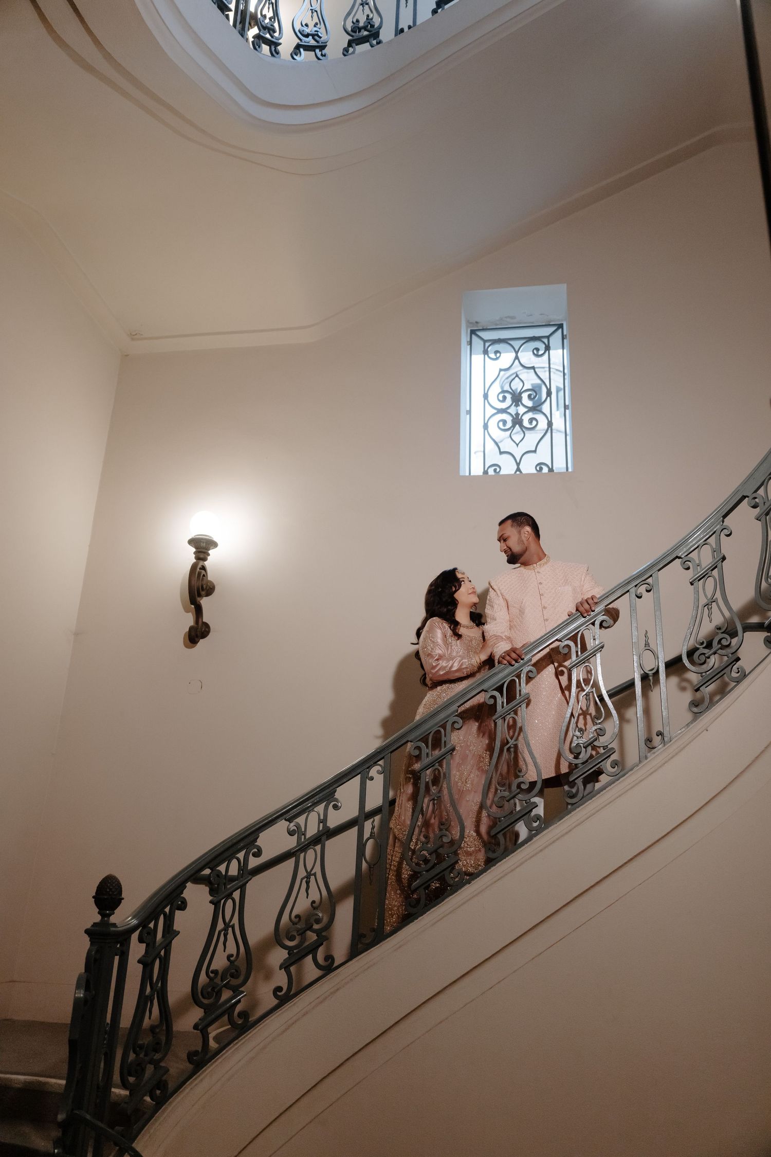 A couple poses on an ornate staircase with decorative iron railings and window light.