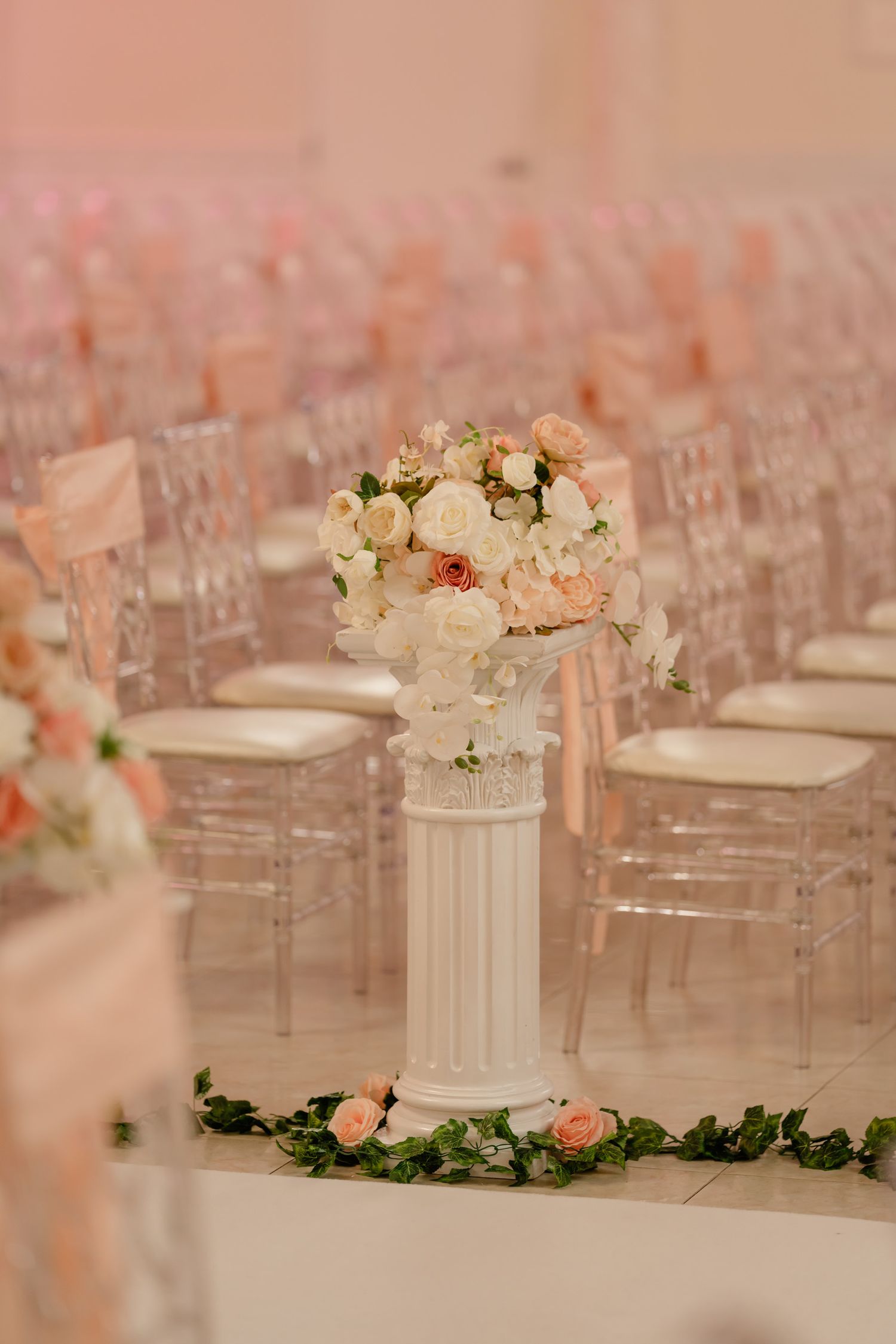 Elegant wedding ceremony setup with white and pink floral arrangements on classical columns and ghost chairs.