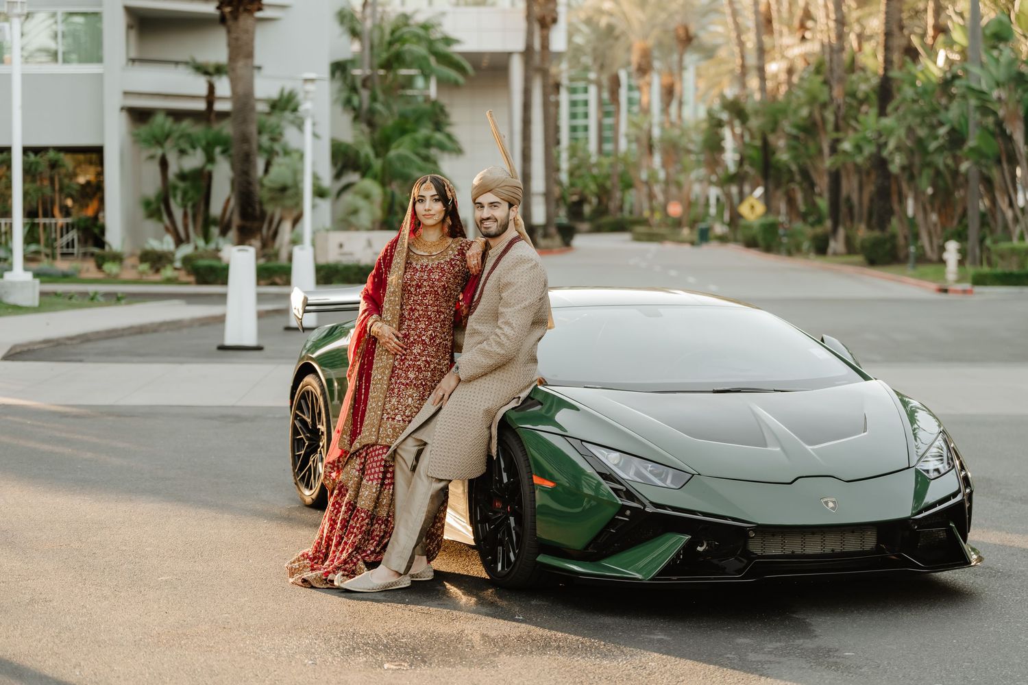 South Asian couple in traditional wedding attire posing with a green Lamborghini Huracan on a palm-lined street.