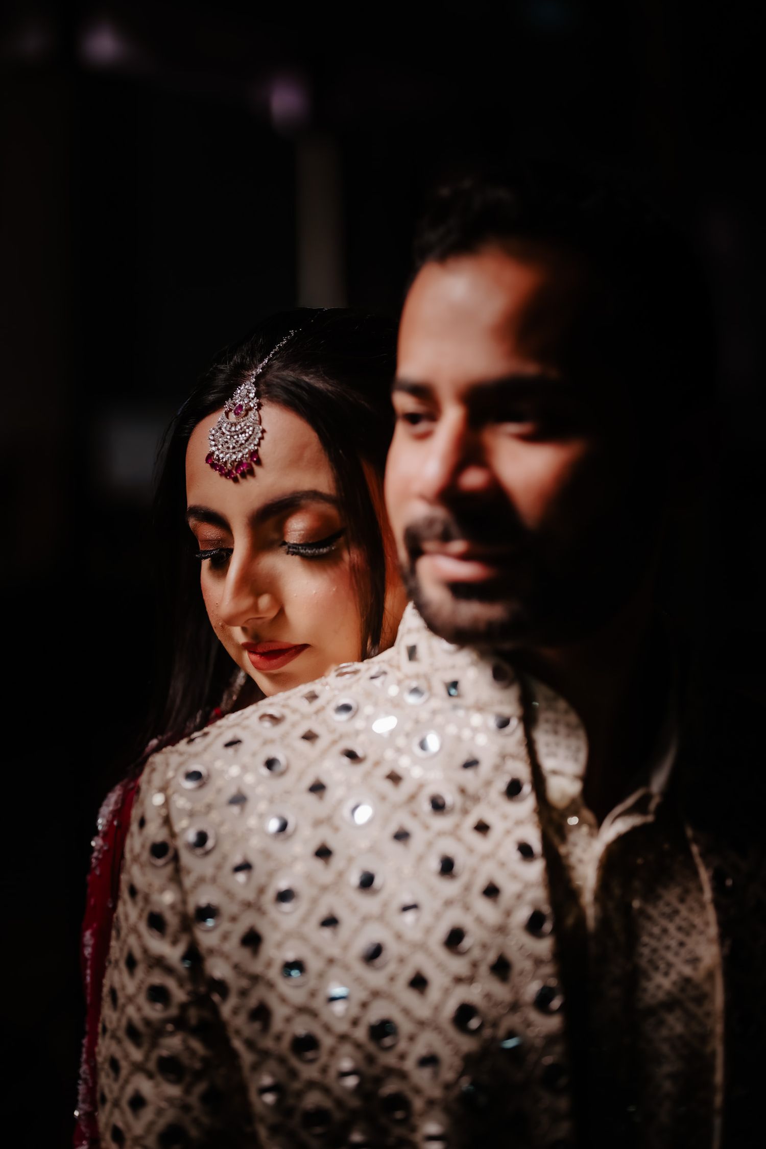 A couple in traditional South Asian wedding attire share an intimate moment in moody lighting.