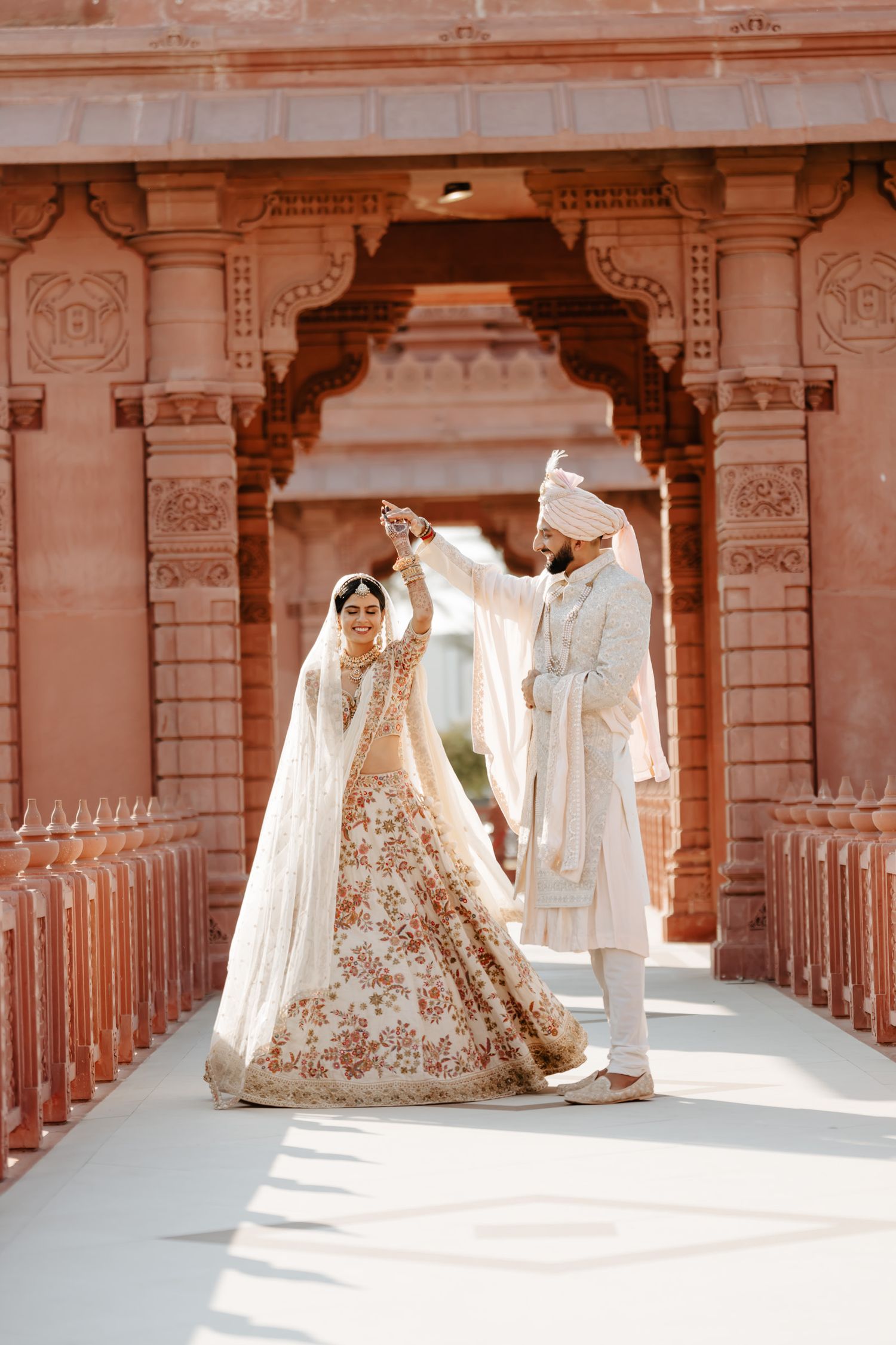 A traditional Indian wedding couple poses under an ornate pink stone archway in elegant ceremonial attire.