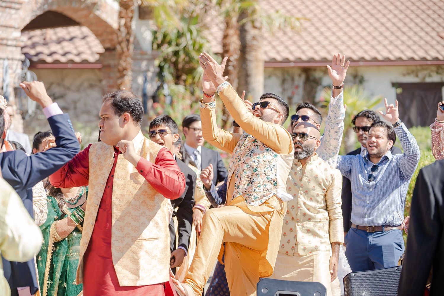 A group of wedding guests raise their arms in celebration during an outdoor reception.