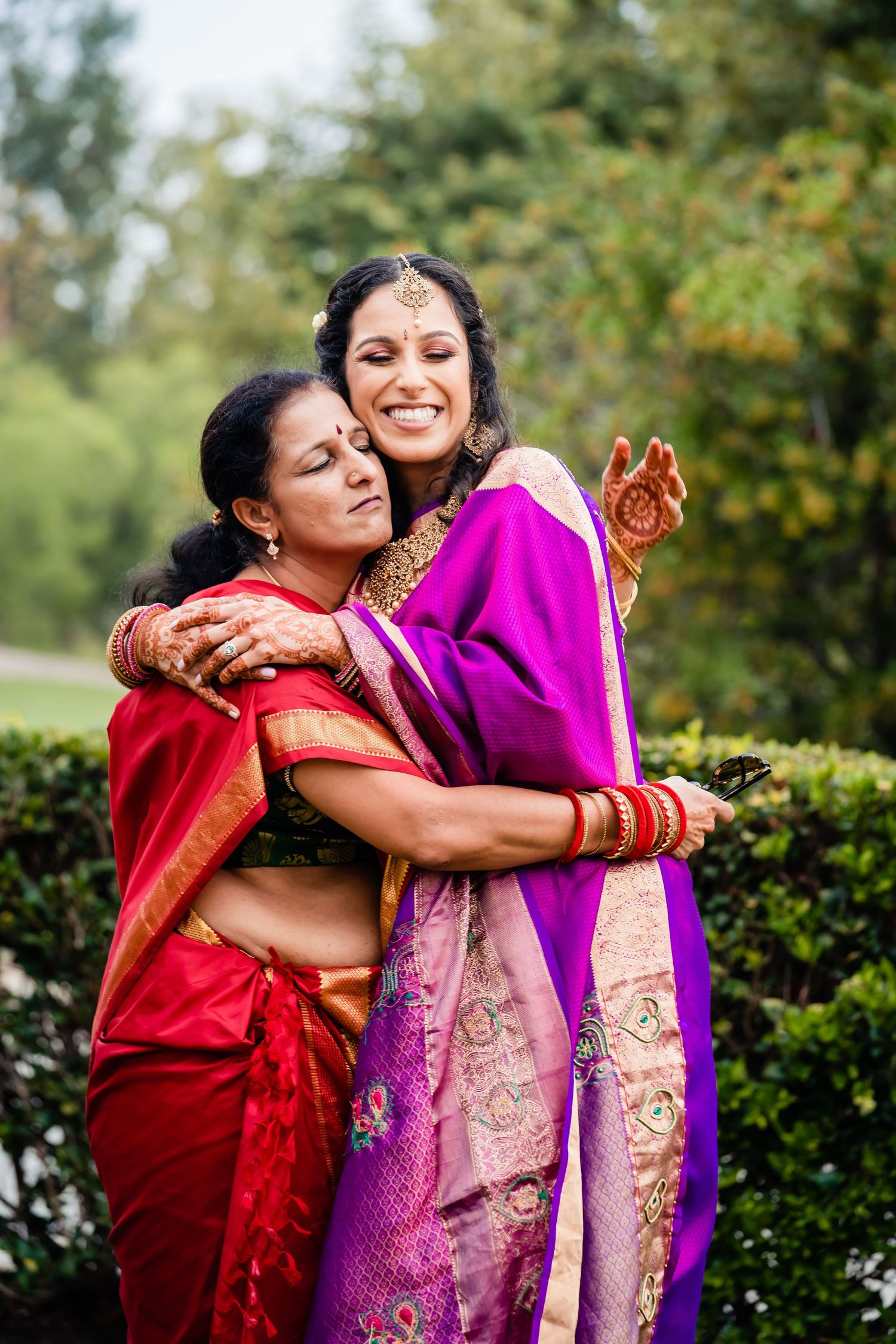 Two people in traditional South Asian attire share joyful embraces in an outdoor garden setting.