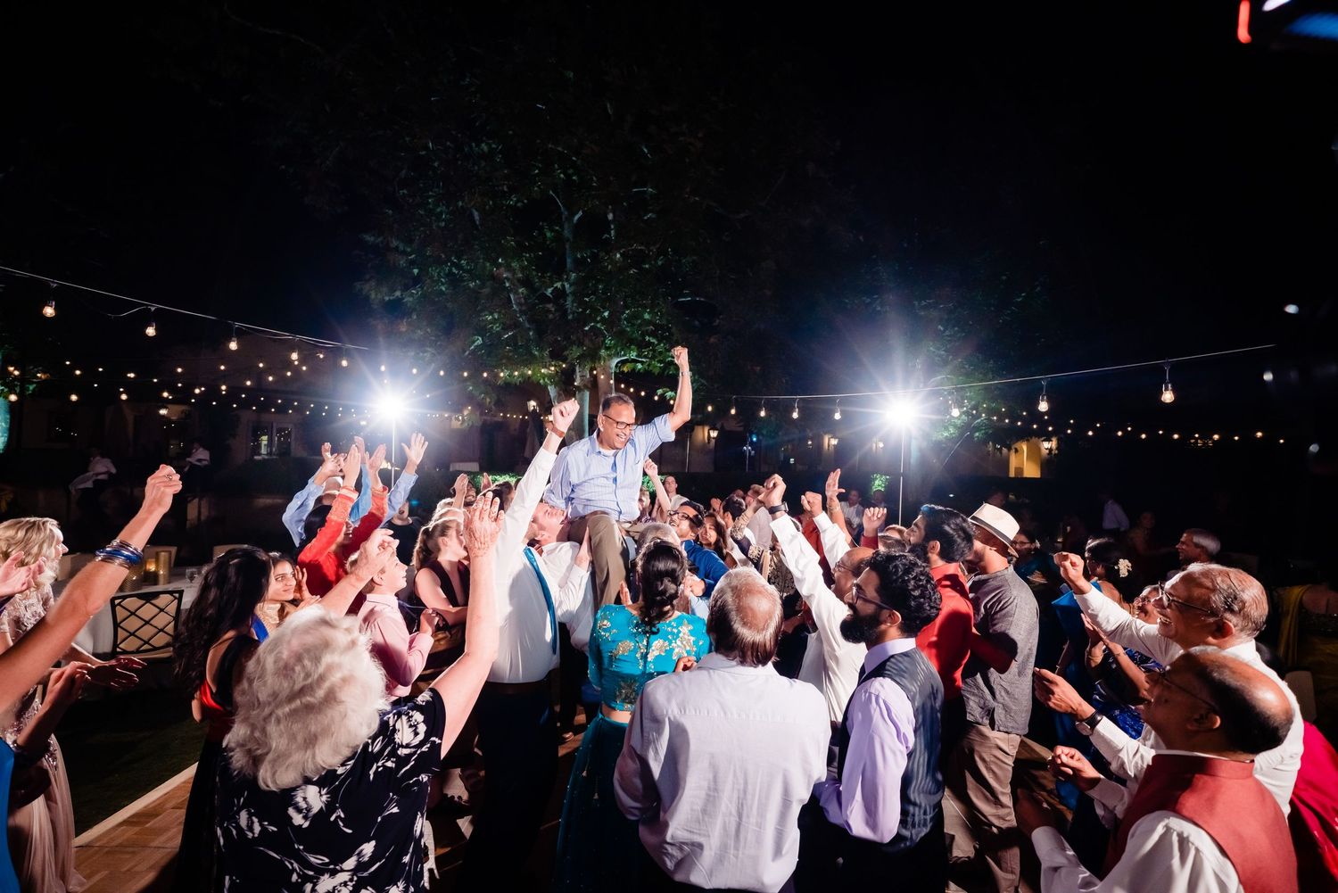 An energetic crowd dances and celebrates at an outdoor nighttime wedding reception with string lights overhead.