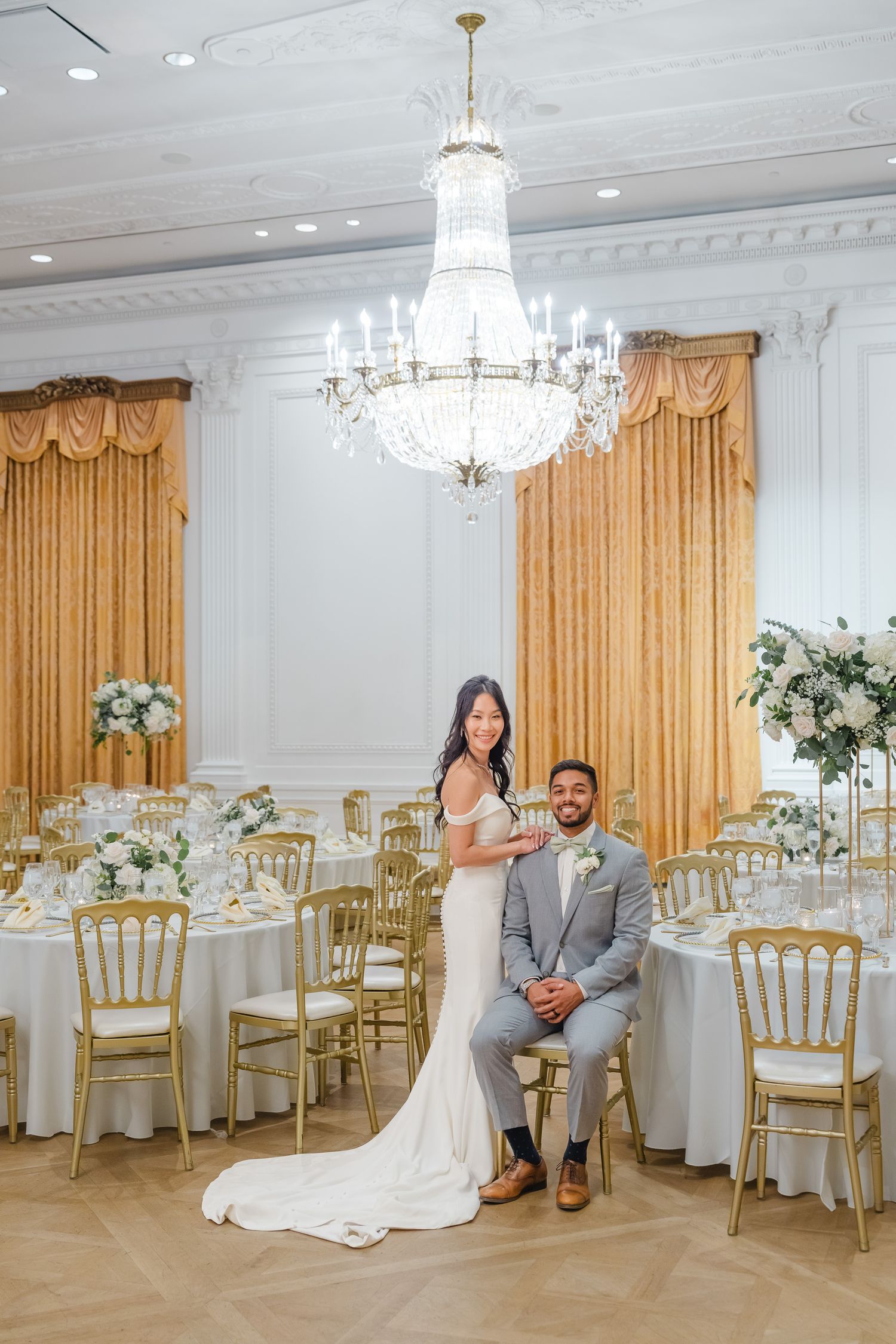 Elegant wedding reception venue with crystal chandeliers, gold chairs, and white floral centerpieces on round tables.