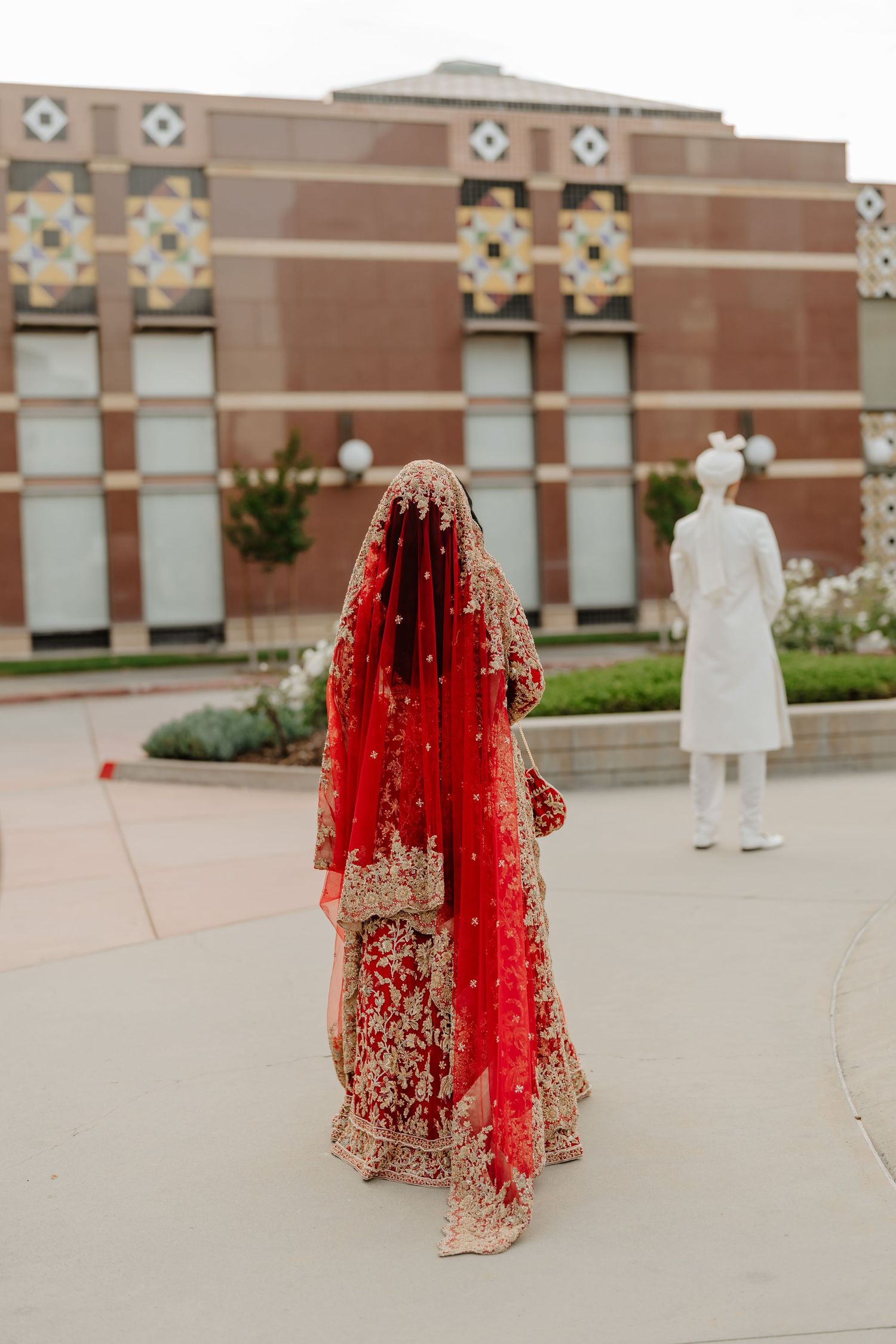 Bride in red and gold bridal lehenga with dupatta veil walks toward groom in white sherwani at outdoor wedding venue.