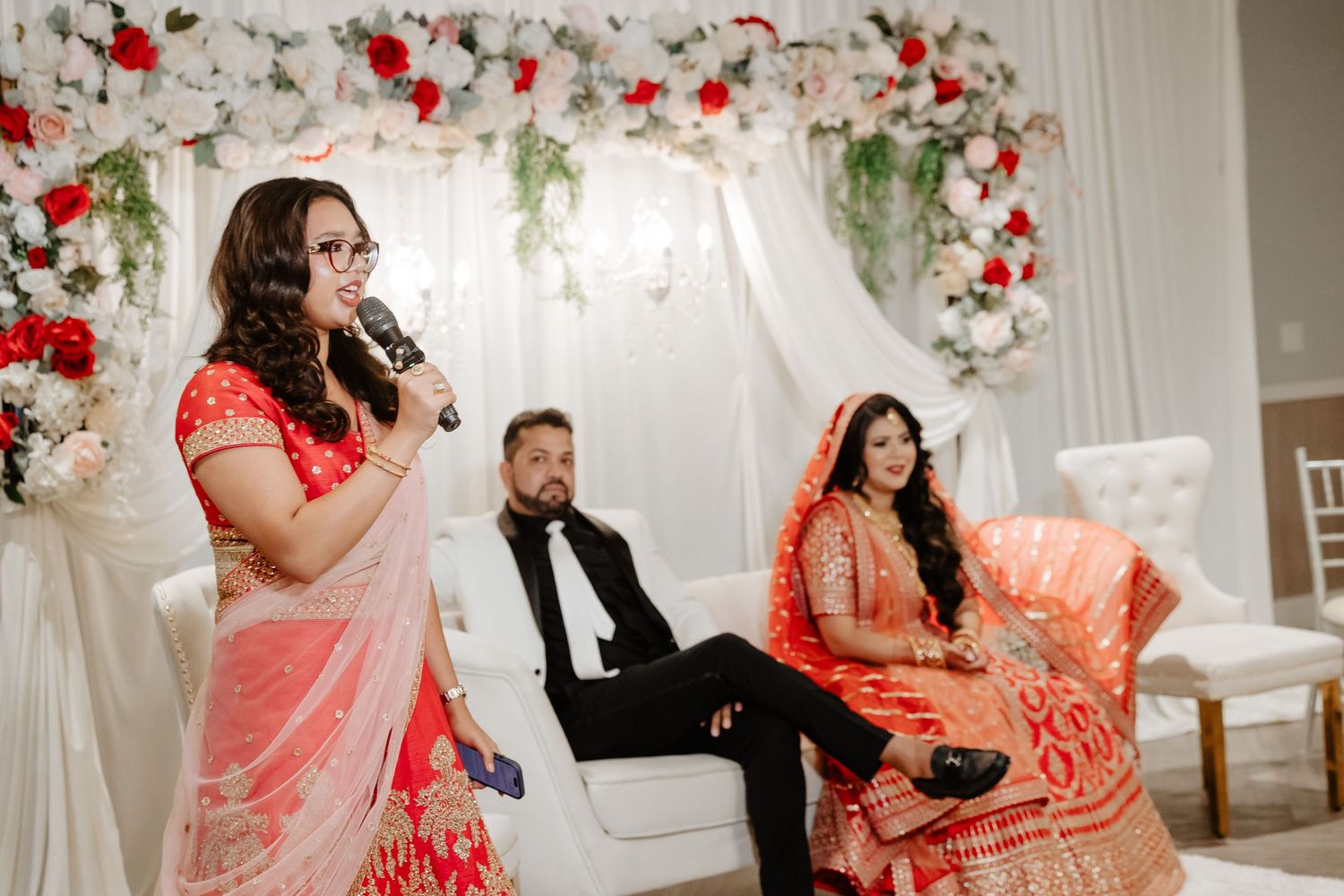 Woman in red saree speaks at mic during Indian wedding ceremony with bride and groom seated behind her.