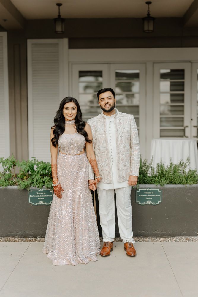 South Asian couple in traditional wedding attire posing together outdoors, woman in pink sequin lehenga, man in ivory sherwani.