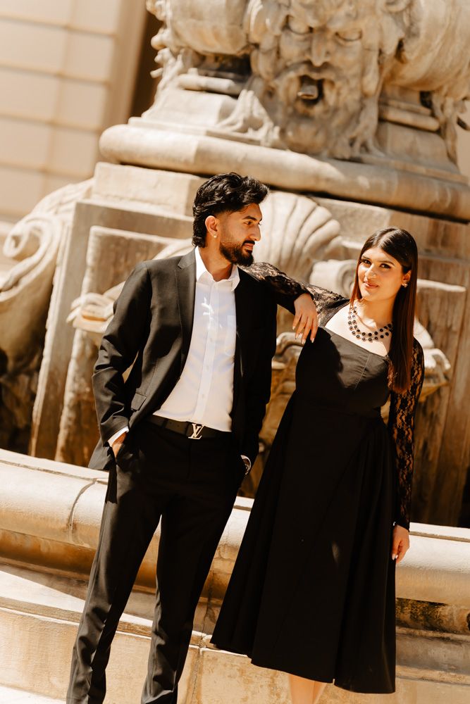 Elegant couple in black formal attire posing by an ornate stone fountain in a classic European setting.