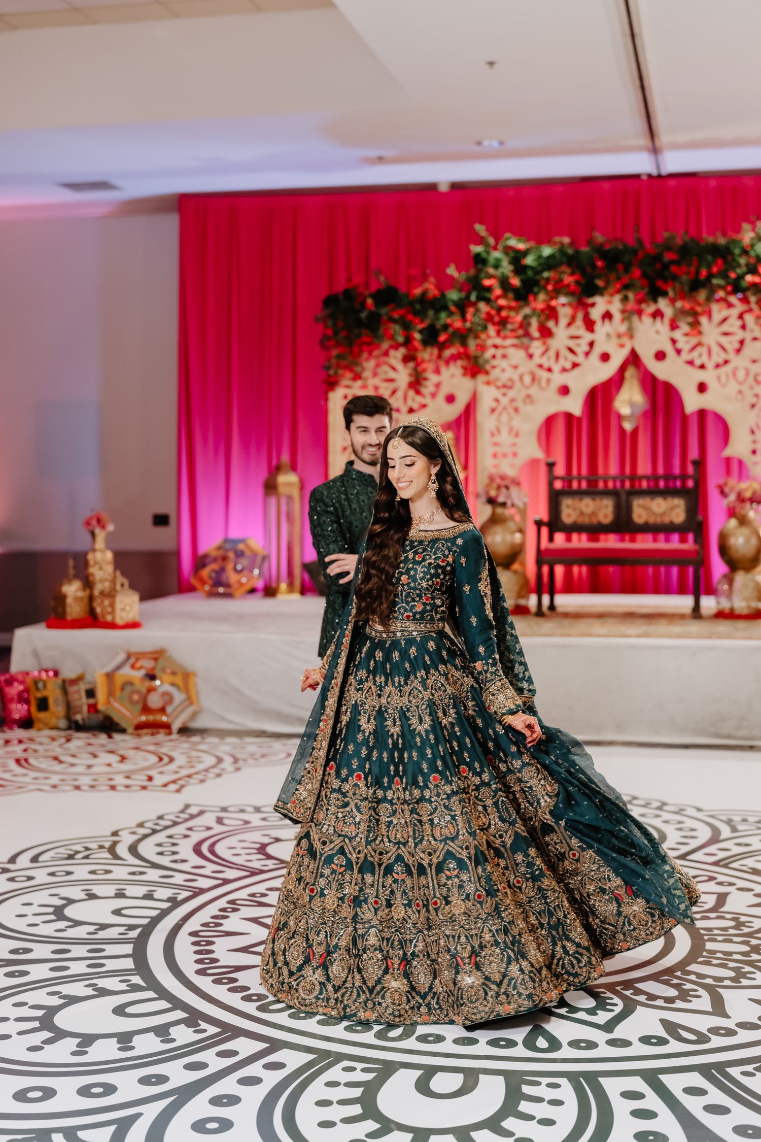 Ornate dark green and gold embroidered lehenga displayed on patterned floor with red backdrop.