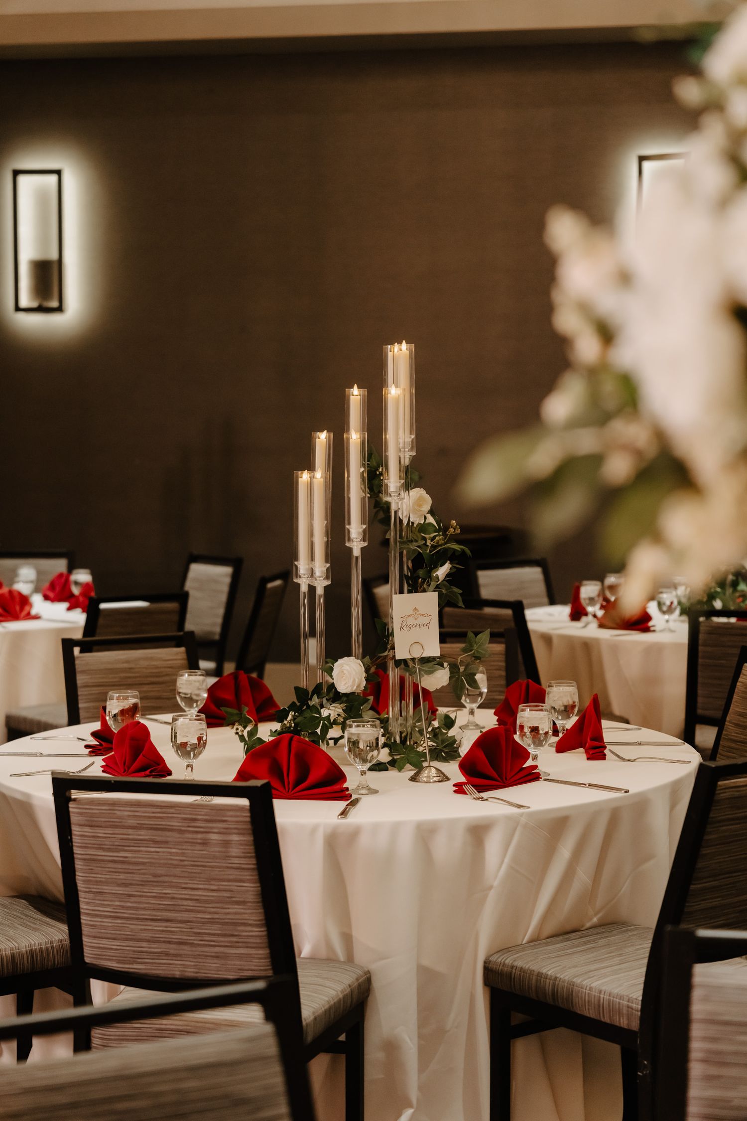 Elegant wedding table setting with tall gold candles, red napkins, and white tablecloth in a dimly lit restaurant.