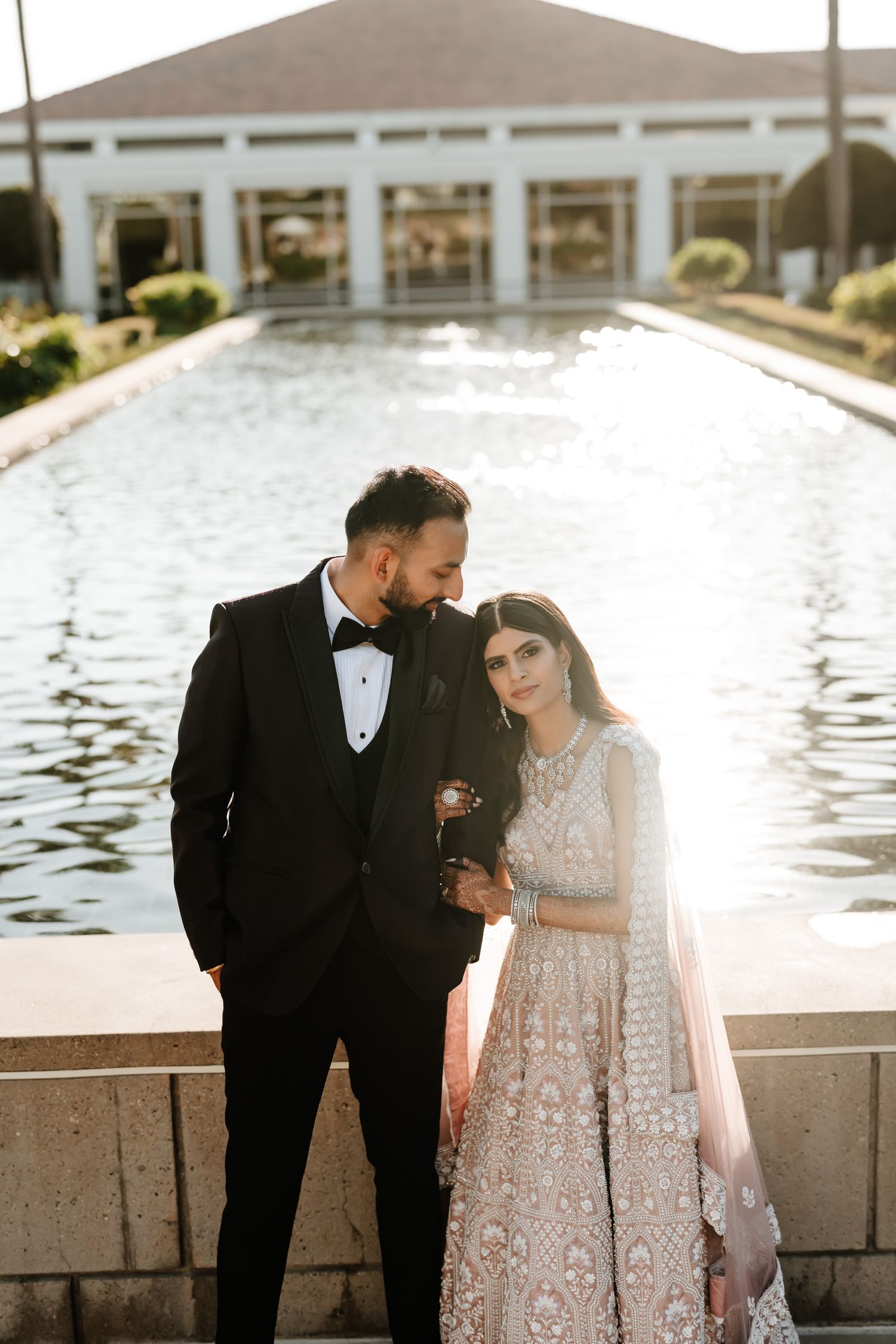 Elegant couple in formal attire posing by reflective pool at sunset with white building in background.