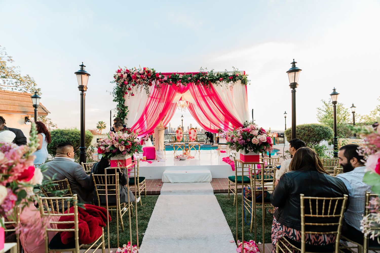 Pink floral wedding ceremony setup with draped mandap and gold chairs on an outdoor terrace.