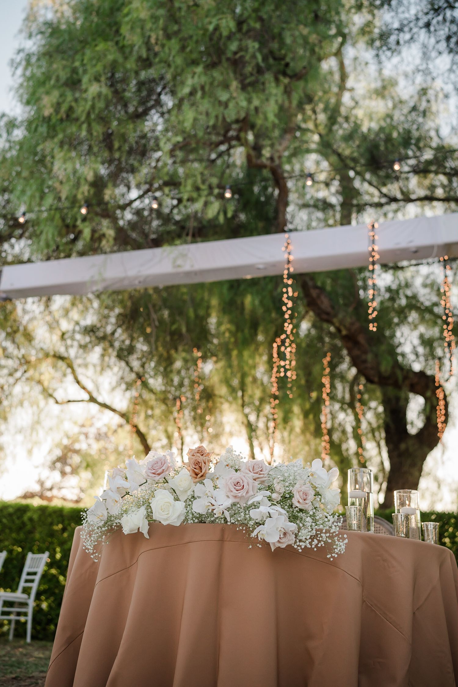 Elegant wedding reception setup with string lights and white floral arrangements under trees at dusk.