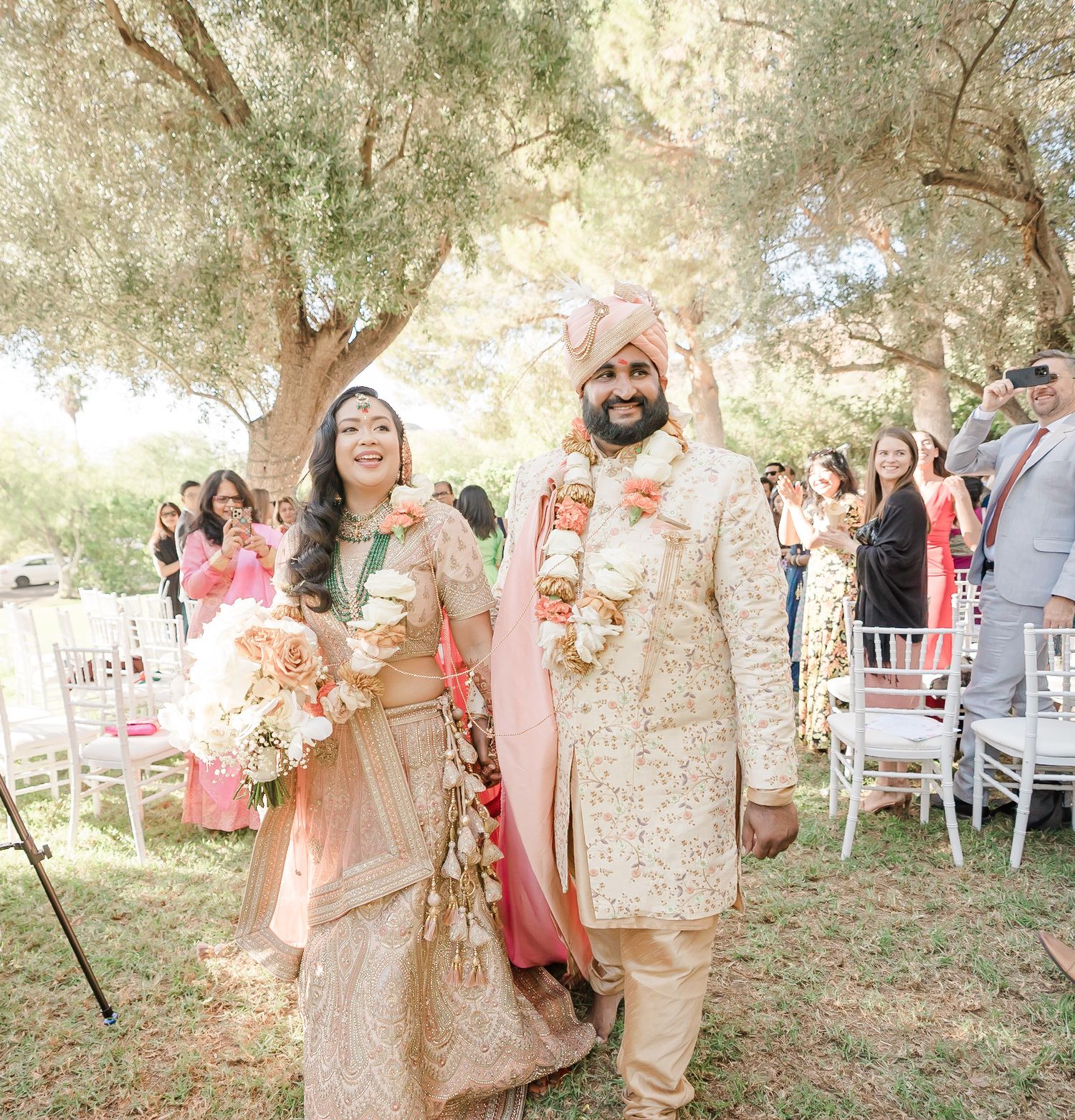 Wedding party gathers in a scenic outdoor setting with pink and coral color scheme under shady trees.