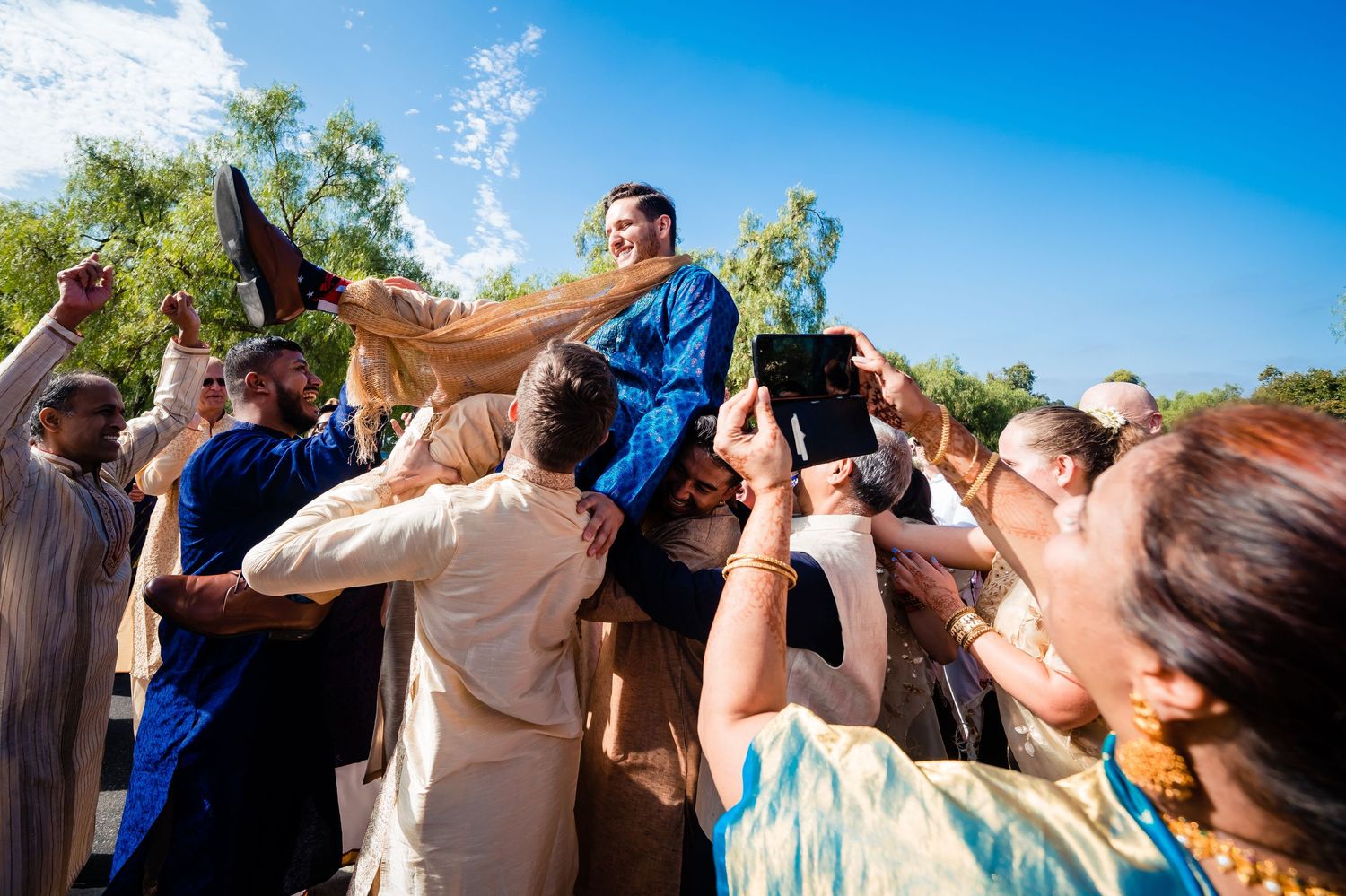A group of people raise their glasses in a celebratory toast against a bright blue sky on a sunny day.