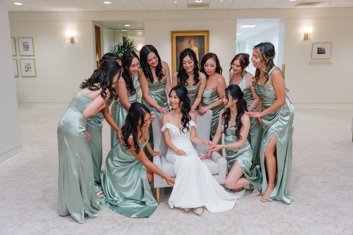 Bridal party in sage green dresses gathered around seated bride in white gown for indoor wedding photo.