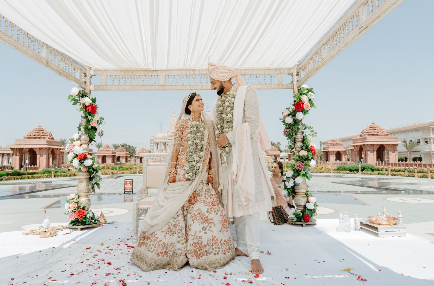 Beachside wedding mandap decorated with white drapes and floral arrangements against a bright blue sky.