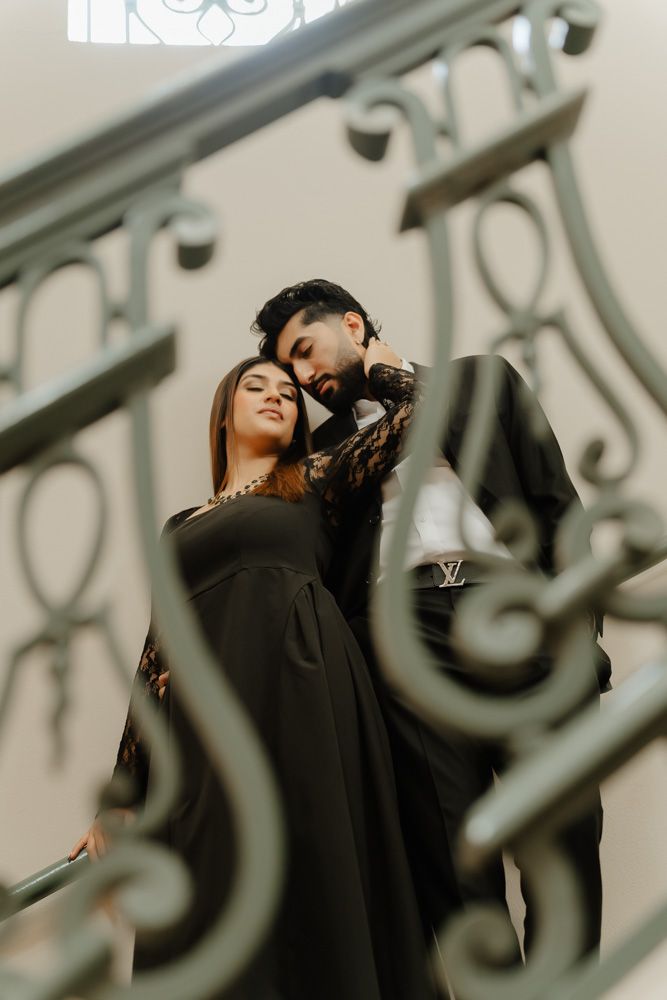 Elegant couple in black outfits posing on ornate staircase, man embracing woman from behind in romantic portrait.
