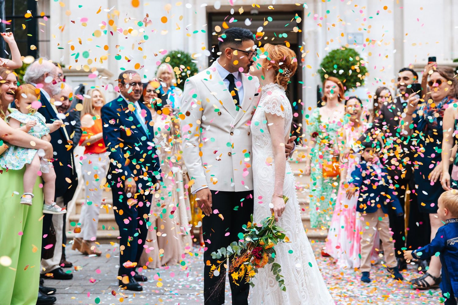 Guests throwing colourful confetti as the couple kisses during the wedding exit in London.