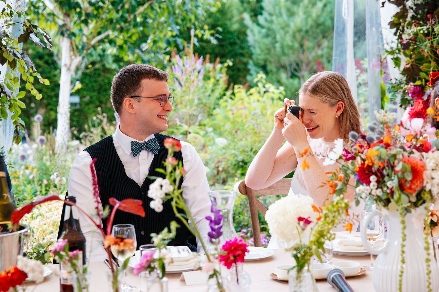 A laughing bride photographs her groom at a colorful outdoor wedding reception table surrounded by vibrant floral arrangements.