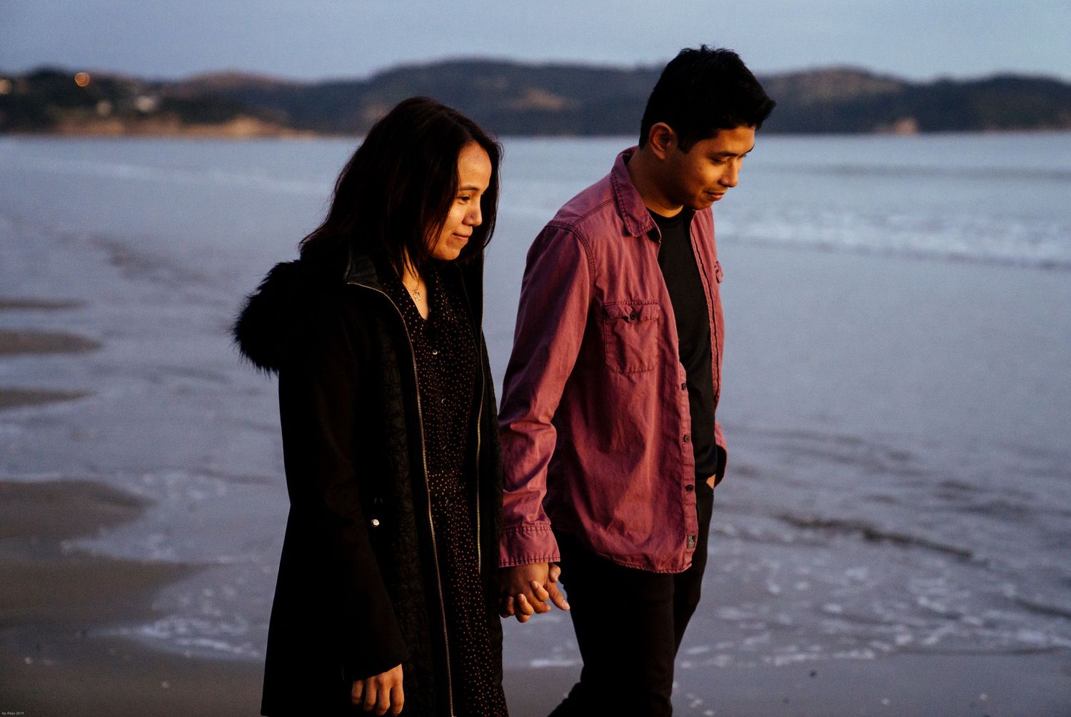 Couple walking along beach shoreline at dusk with mountains in background.