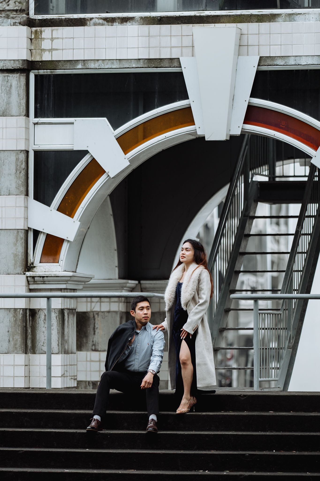 Two people pose under white arched architectural features in an urban setting.