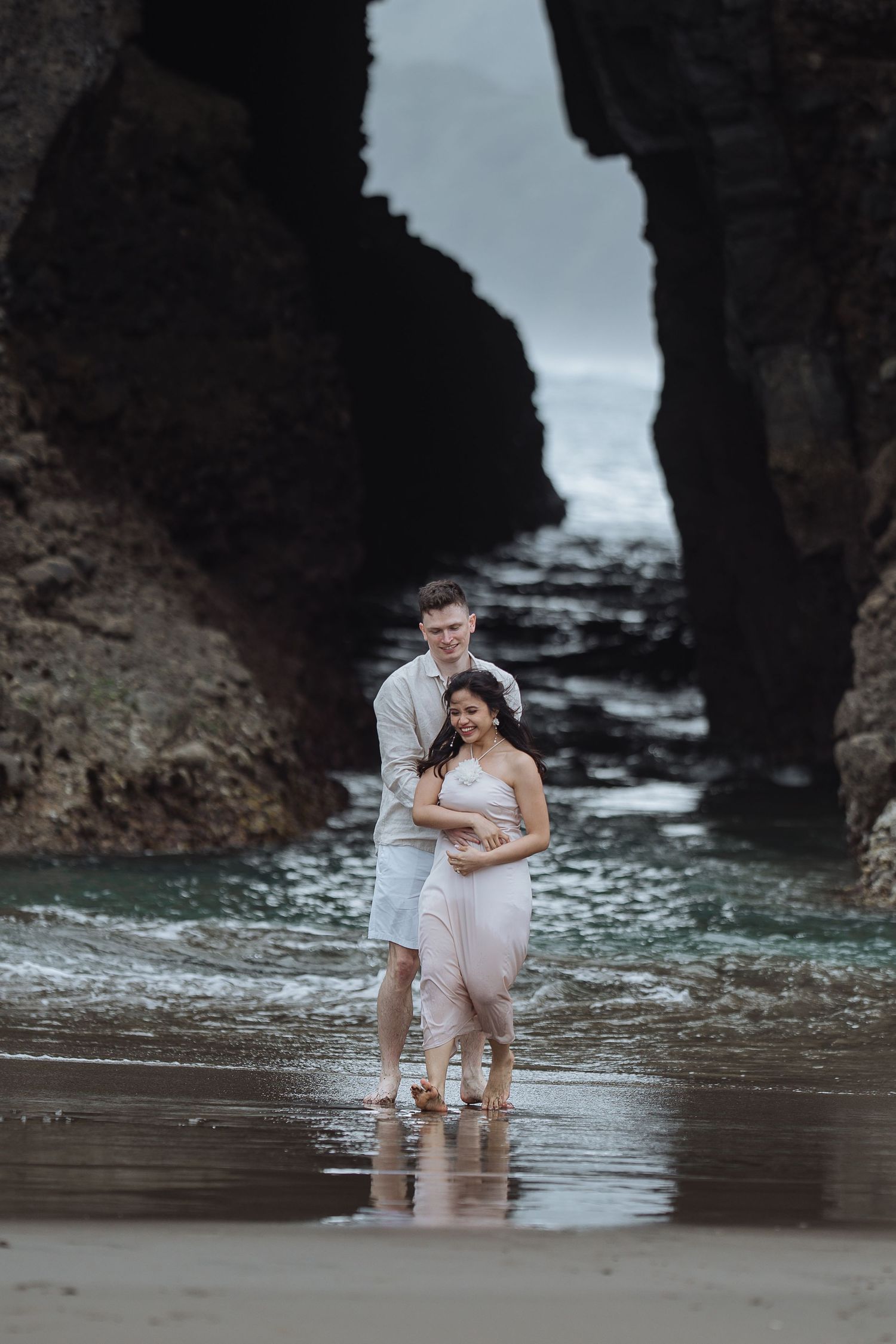 Couple embracing under natural rock arch formation on beach with waves visible.