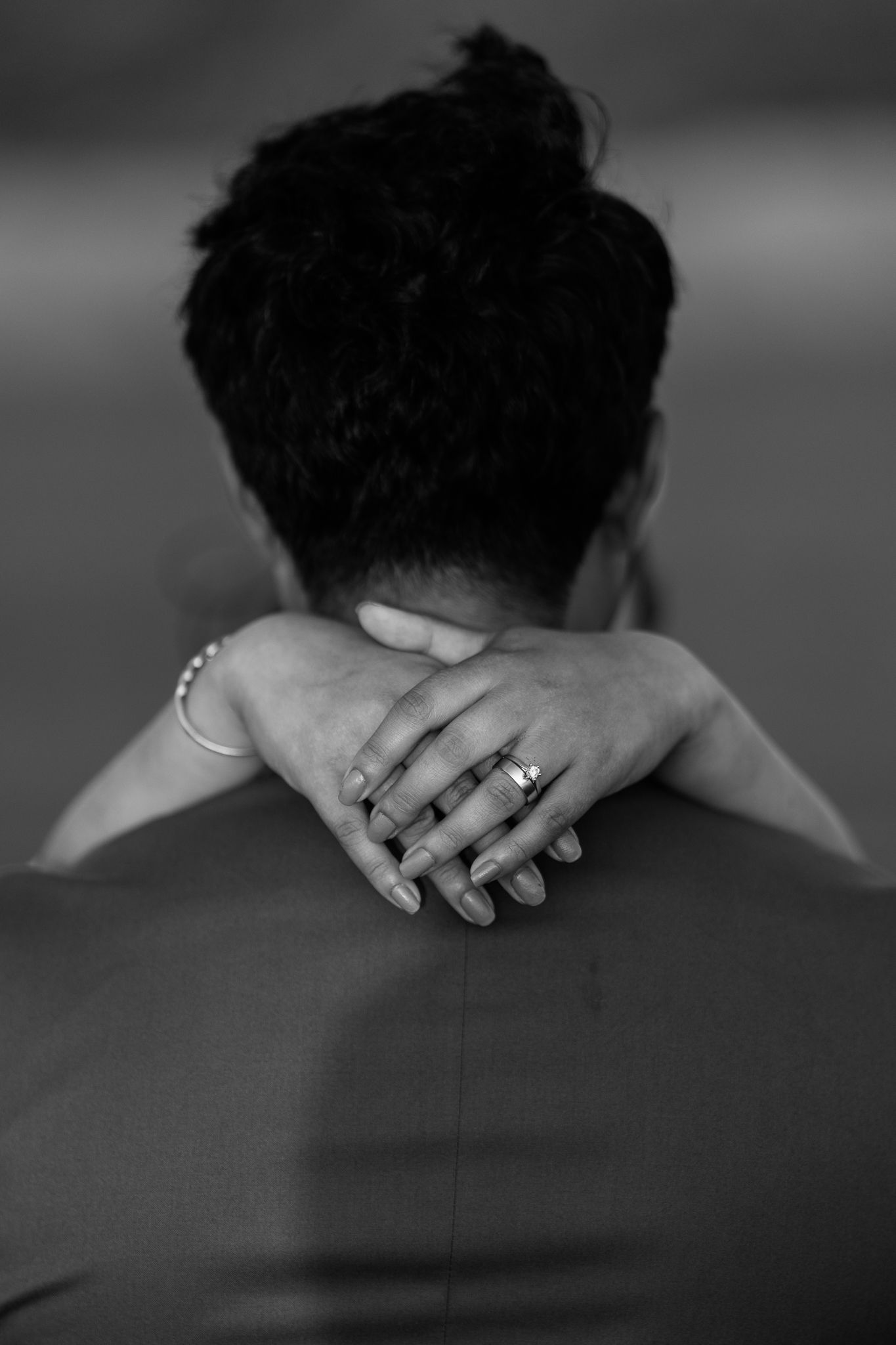 A close-up black and white photo showing hands resting on shoulders with wedding rings.