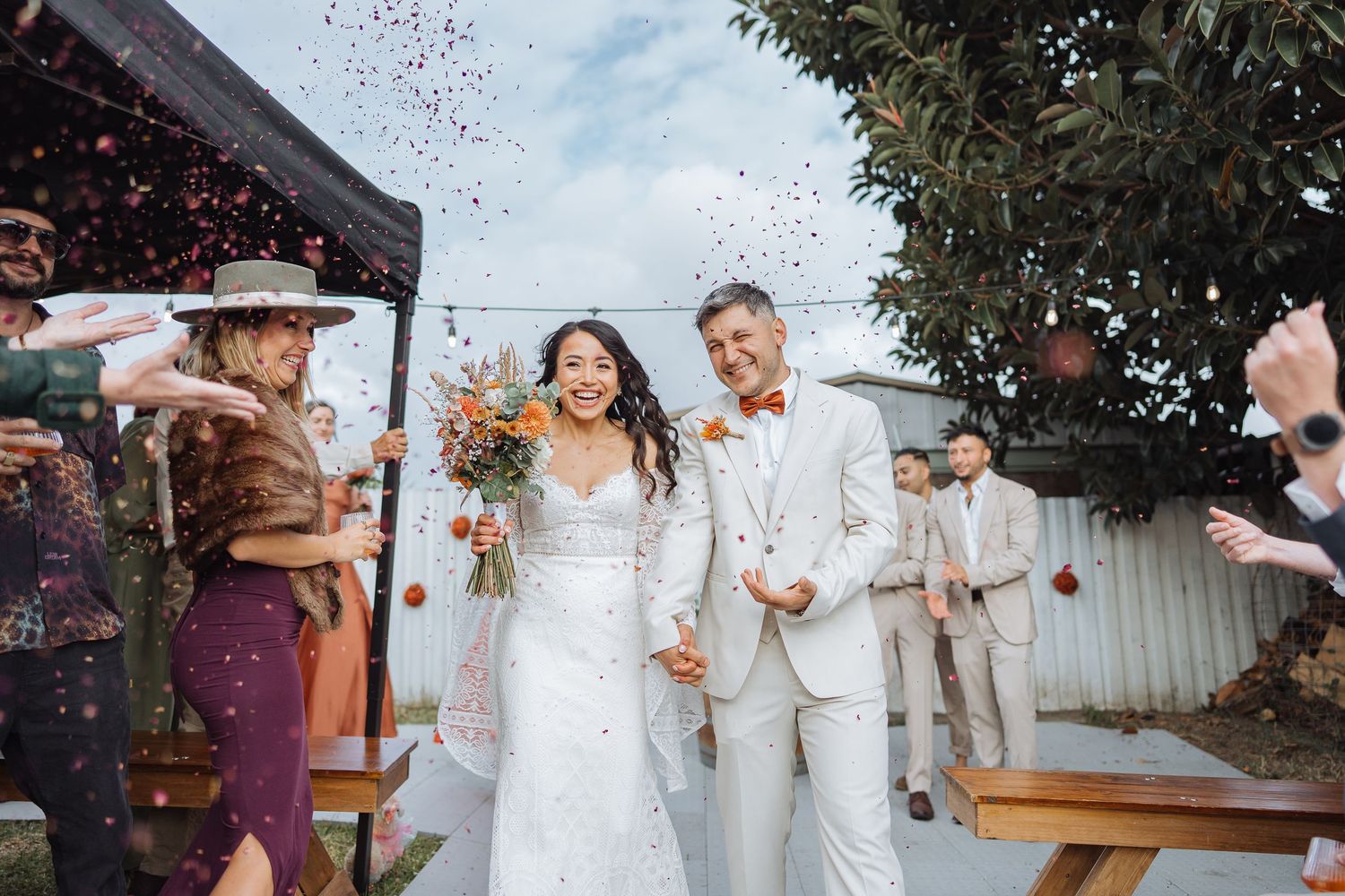 Bride and groom walk down aisle as guests toss colorful confetti at outdoor wedding ceremony.