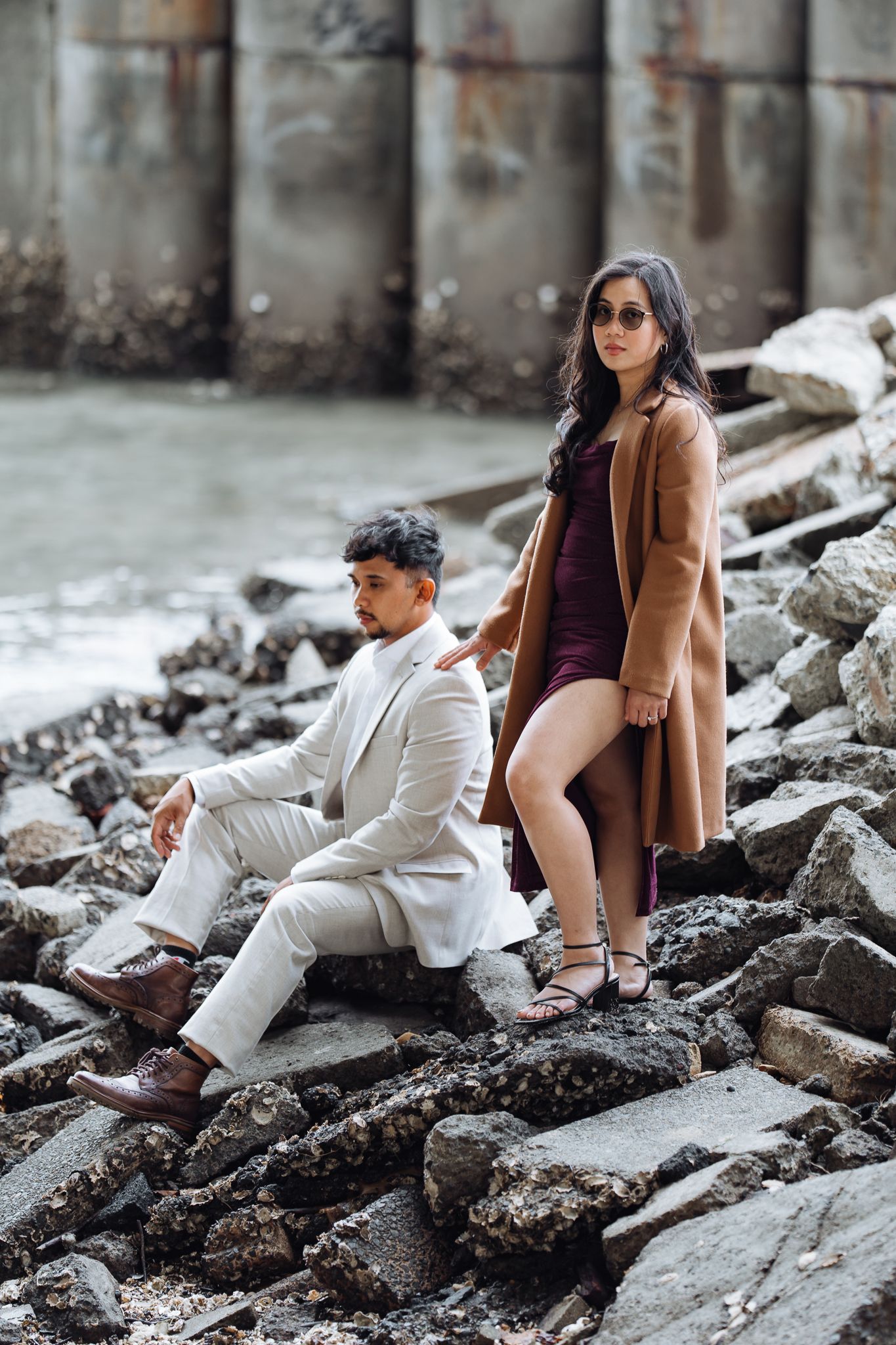 Stylish couple in contrasting outfits posing on rocky waterfront terrain.