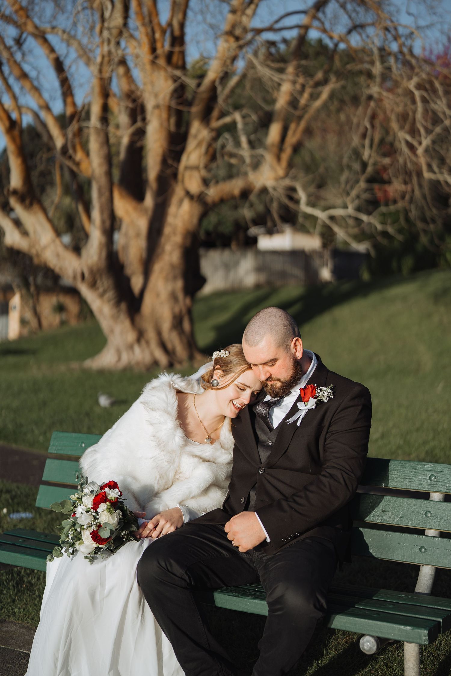 Newlyweds share intimate moment on park bench surrounded by winter trees.
